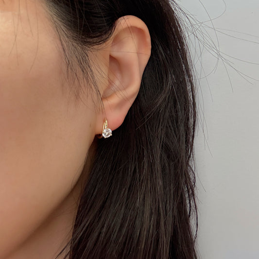 Close-up of an ear wearing a gold earring with a clear gemstone against a neutral background