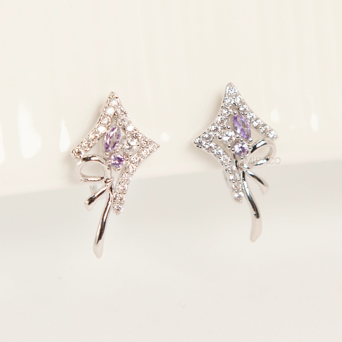 Pair of silver-tone bow-shaped clip-on earrings featuring purple amethyst stones, pave crystals, and comfortable invisible coil back closures.