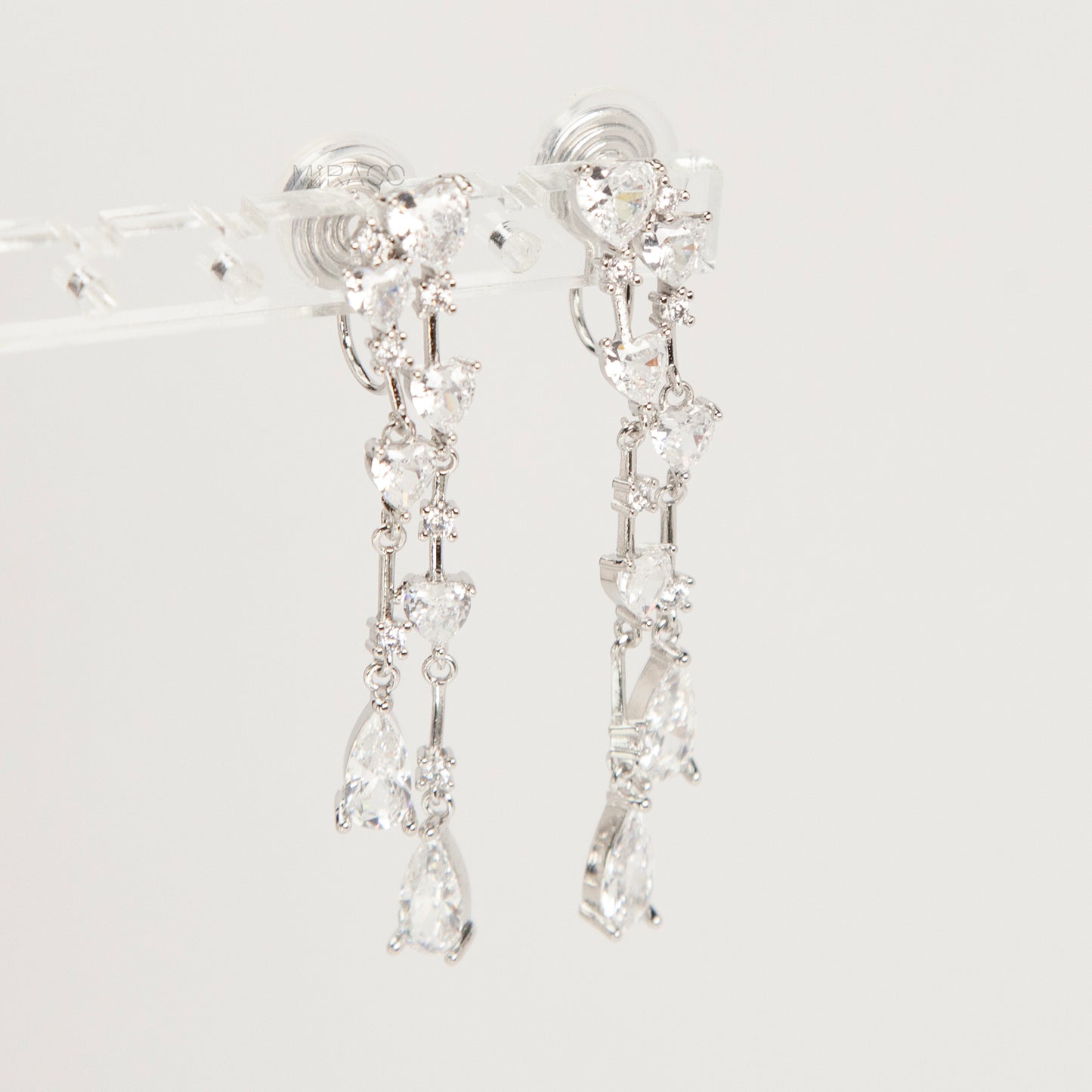 Pair of silver-toned cascading clip-on earrings featuring heart-shaped and teardrop cubic zirconia crystals on a dainty drop chain with a comfortable coil back.