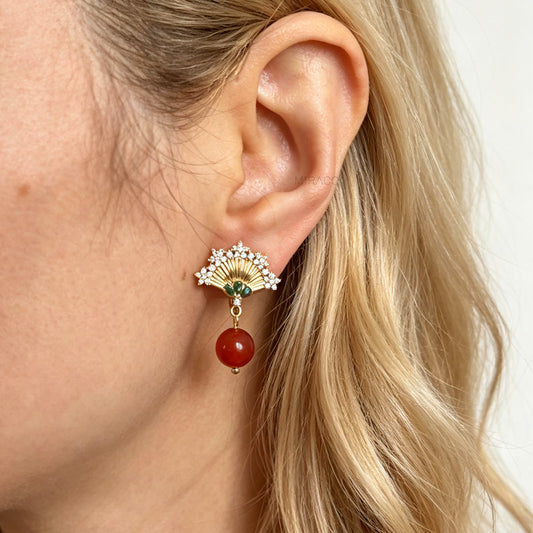 Gold Art Deco fan-shaped clip-on earrings with green crystal accents, pavé CZ trim, and hanging red agate bead drops worn on an ear model