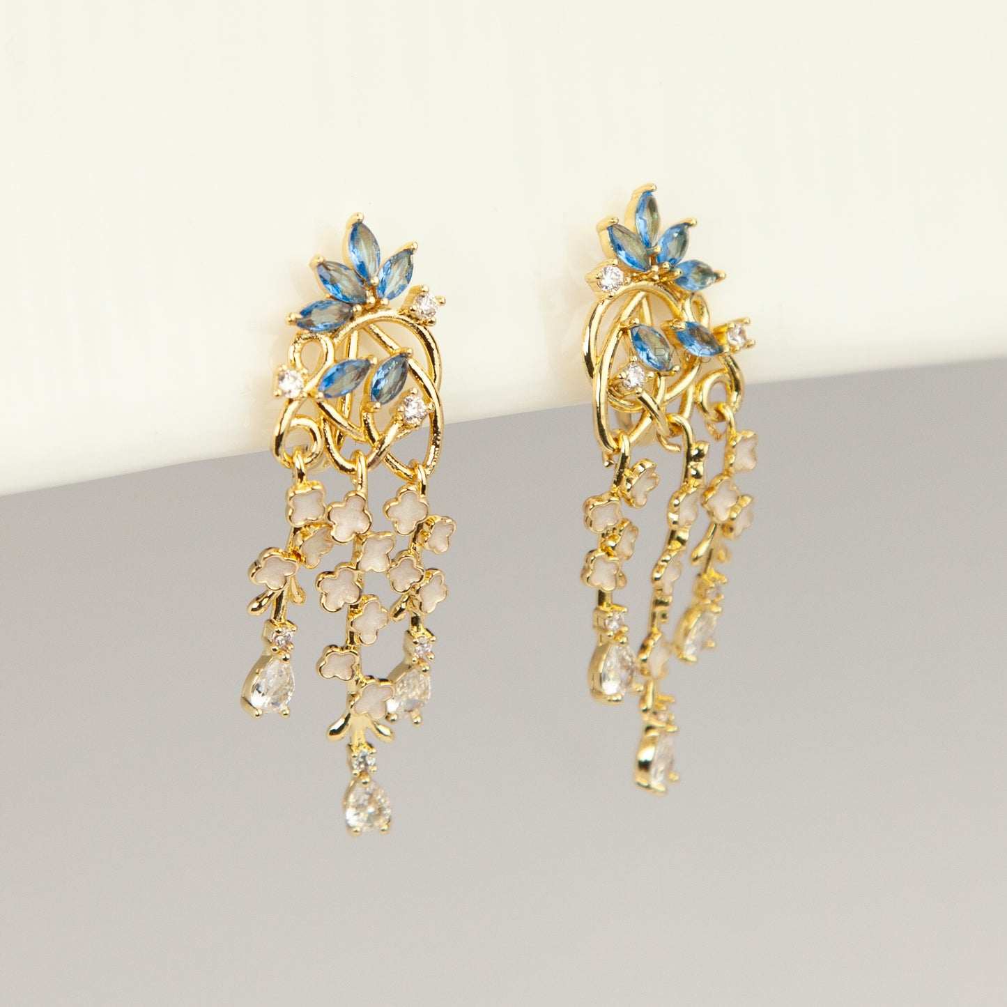 Gold coil-back clip-on chandelier earrings featuring blue marquise petals and cascading white flower dangles with teardrop crystals