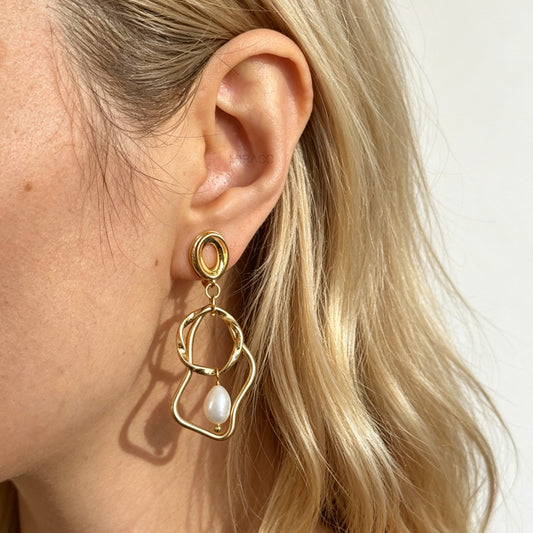 Gold coil-back clip-on statement earrings featuring abstract wavy geometric hoops and dangling teardrop pearl accents.