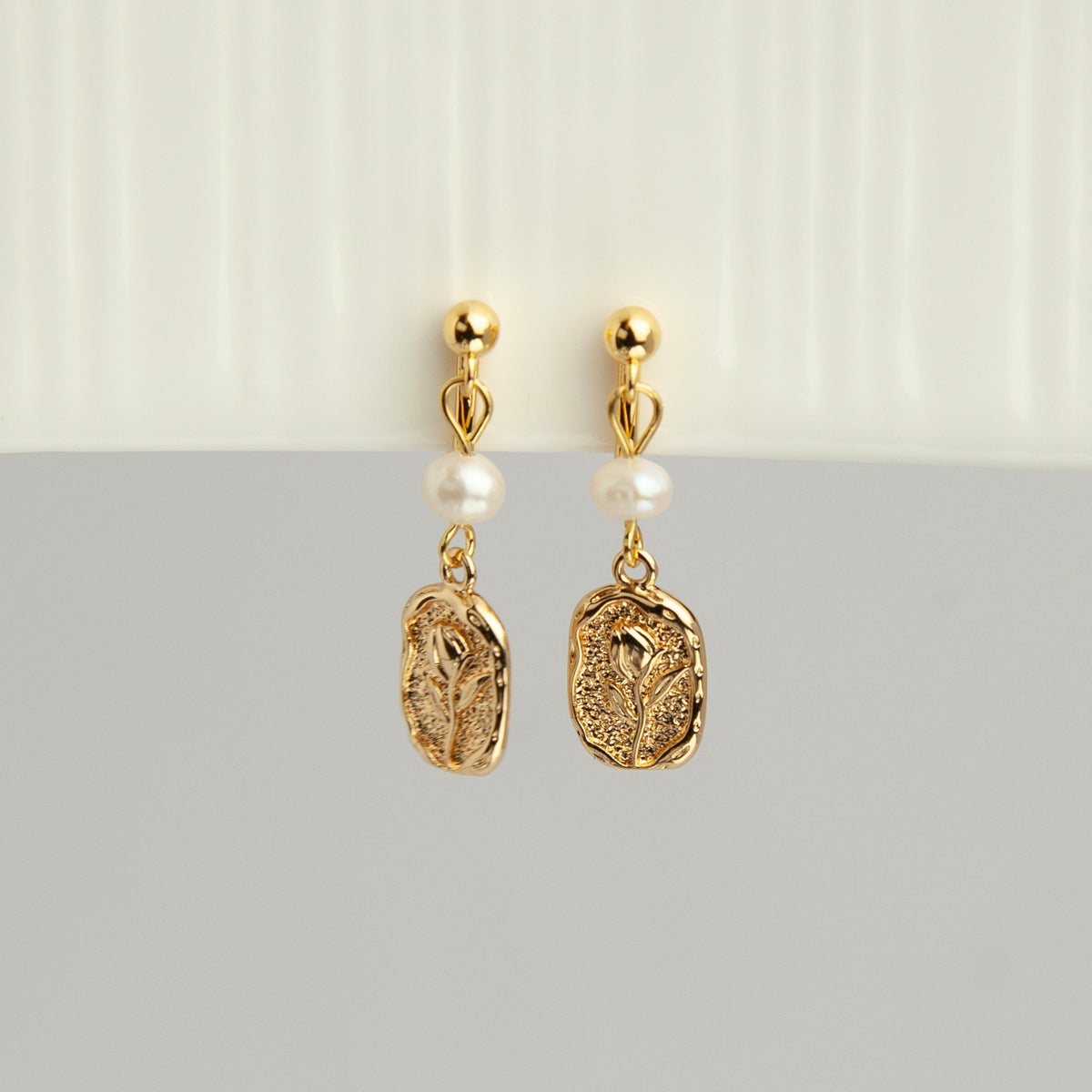 Gold screw-back clip-on drop earrings featuring a lustrous white pearl and a textured oval medallion with an embossed floral design