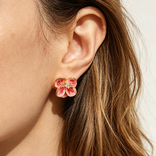 A close-up of a woman's ear wearing a small flower-shaped stud earring with pink petals, gold outlines, and a tiny clear center stone.