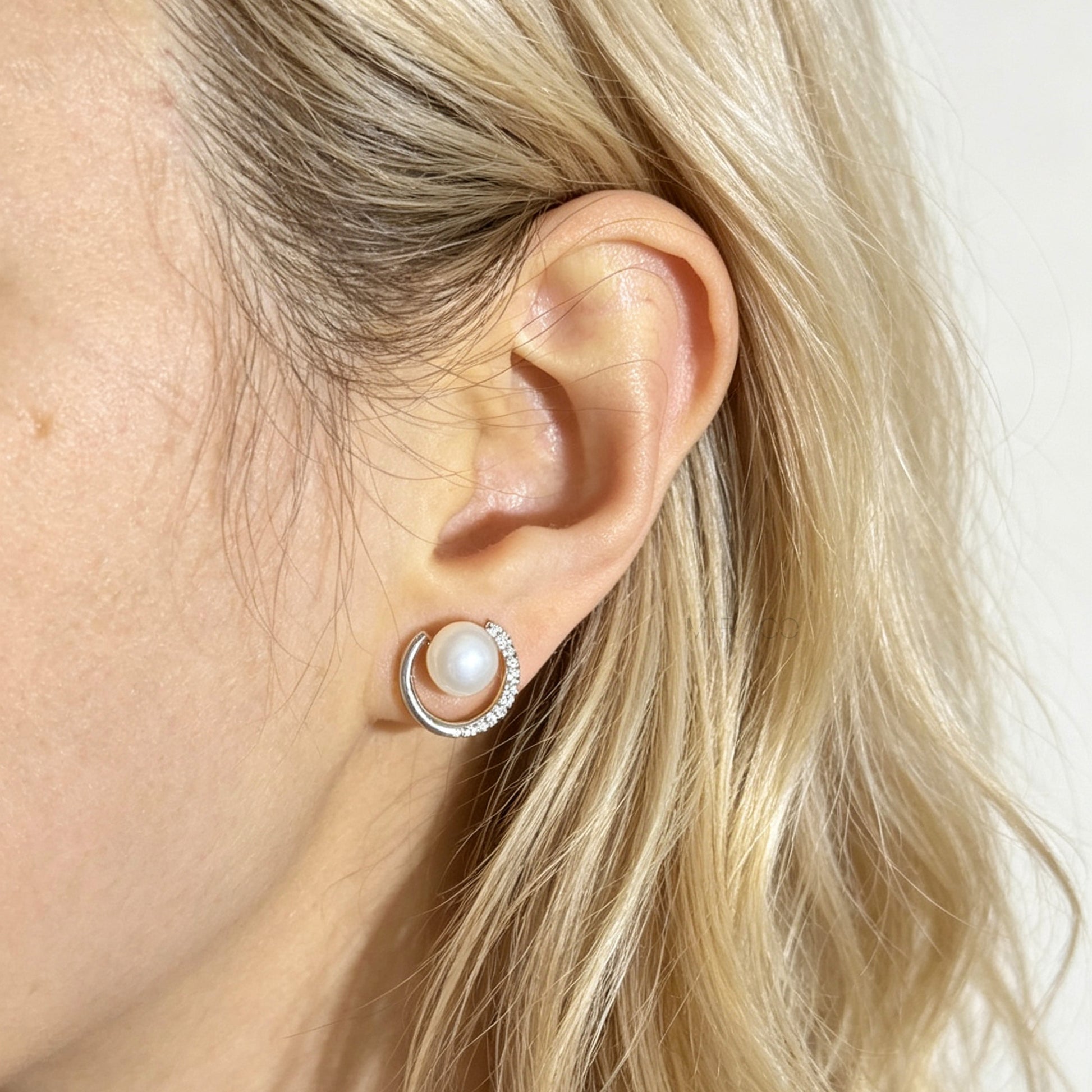 A close-up of a blonde woman's ear wearing a small hoop stud earring with a white pearl and a halo of clear stones.
