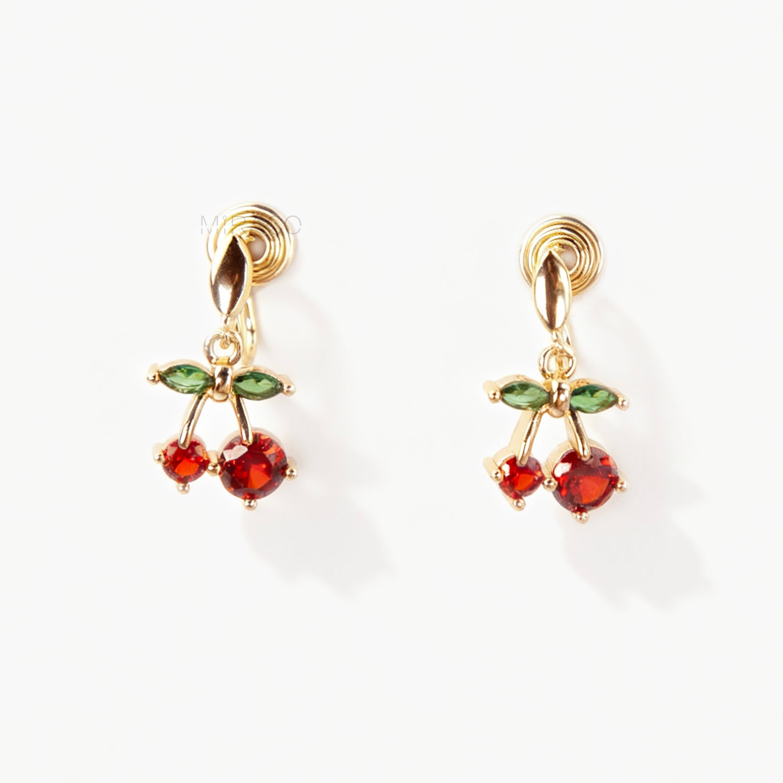Pair of gold-plated coil back clip-on earrings featuring a double-cherry dangle design. The cherries are made of faceted red cubic zirconia stones with green marquise-cut CZ leaves above them. The back is a round, spiraled coil for comfort.