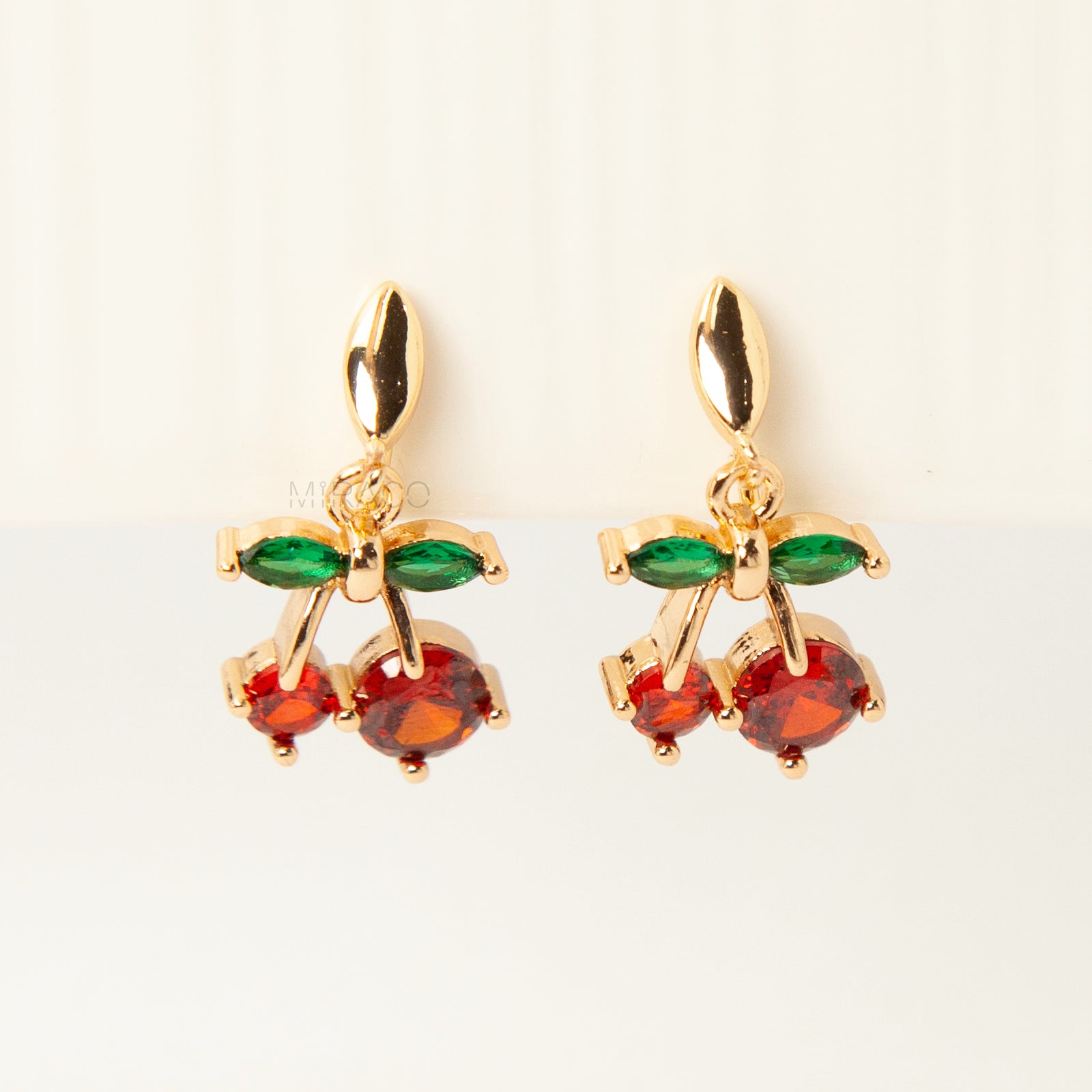 Pair of gold-plated coil back clip-on earrings featuring a double-cherry dangle design. The cherries are made of faceted red cubic zirconia stones with green marquise-cut CZ leaves above them. The back is a round, spiraled coil for comfort.