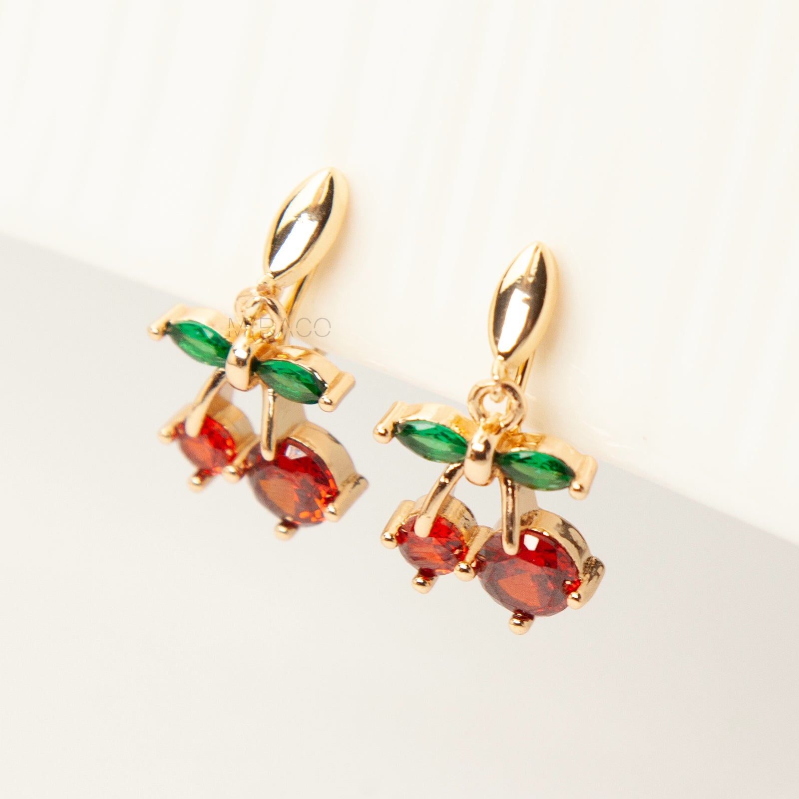 Pair of gold-plated coil back clip-on earrings featuring a double-cherry dangle design. The cherries are made of faceted red cubic zirconia stones with green marquise-cut CZ leaves above them. The back is a round, spiraled coil for comfort.