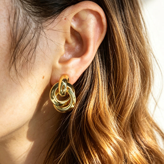 A pair of high-polish gold clip-on earrings featuring three interlocking, dimensional smooth metal hoops arranged in a interlocking knot pattern, attached to clear-padded adjustable metal coil backs.
