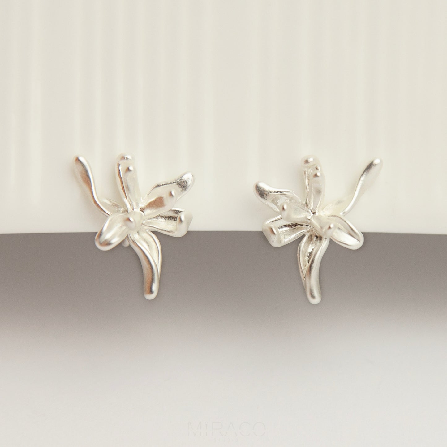 A pair of elegant matte silver floral clip-on earrings from clip-on-earrings-clip216-7.jpg, showcasing an abstract 3D orchid flower design with organic, curved metallic petals and fitted with comfortable, invisible-style adjustable coil backs for non-pierced ears.