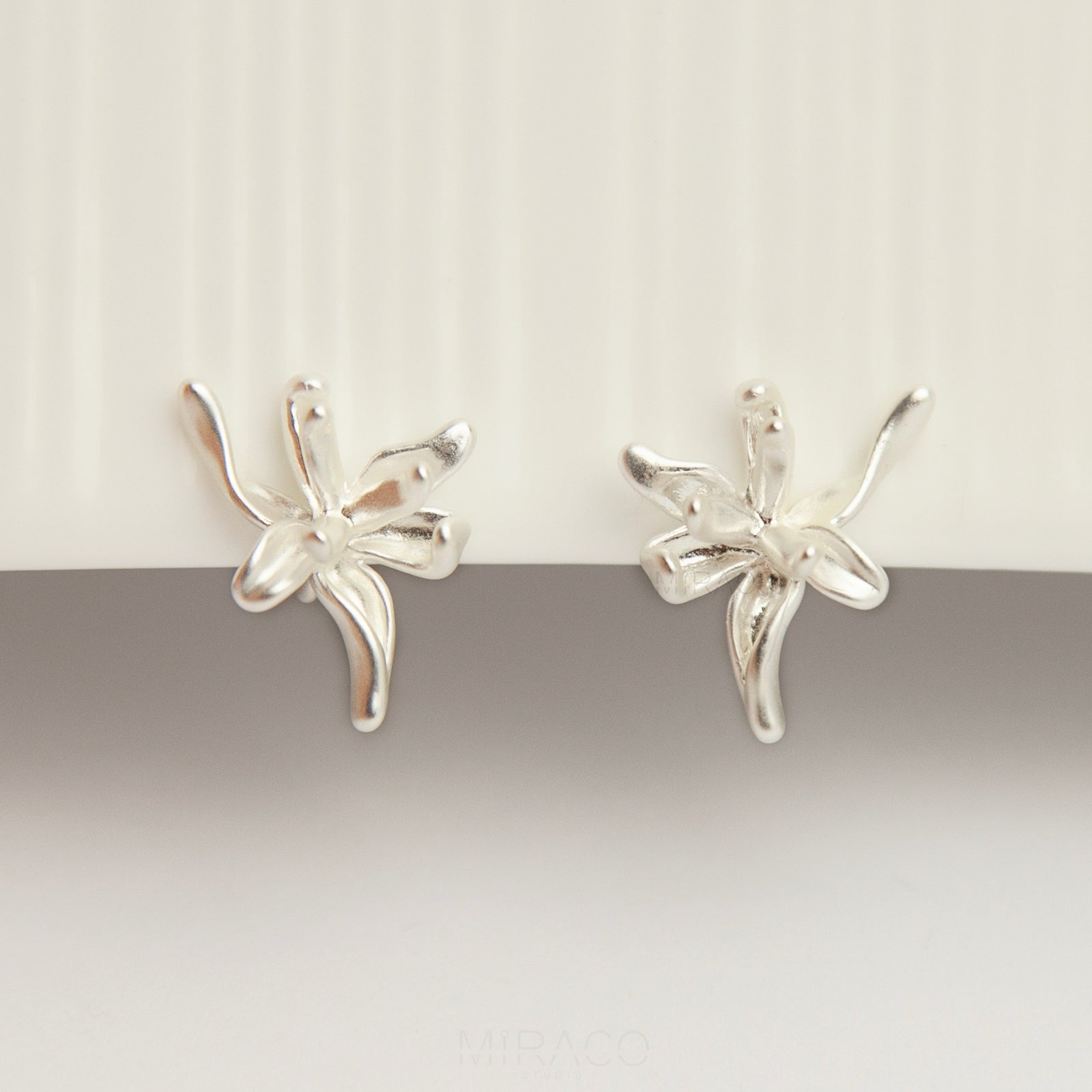 A pair of elegant matte silver floral clip-on earrings from clip-on-earrings-clip216-7.jpg, showcasing an abstract 3D orchid flower design with organic, curved metallic petals and fitted with comfortable, invisible-style adjustable coil backs for non-pierced ears.