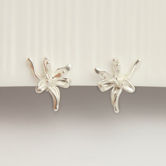 A pair of elegant matte silver floral clip-on earrings from clip-on-earrings-clip216-7.jpg, showcasing an abstract 3D orchid flower design with organic, curved metallic petals and fitted with comfortable, invisible-style adjustable coil backs for non-pierced ears.