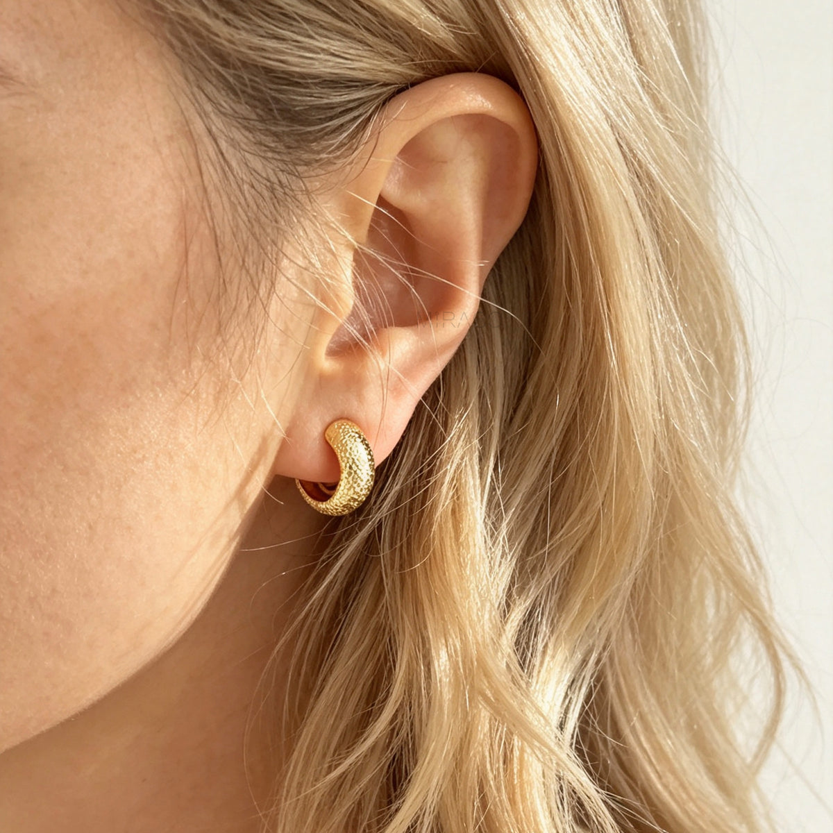 A close-up of a model's ear wearing a textured gold  hoop earring.