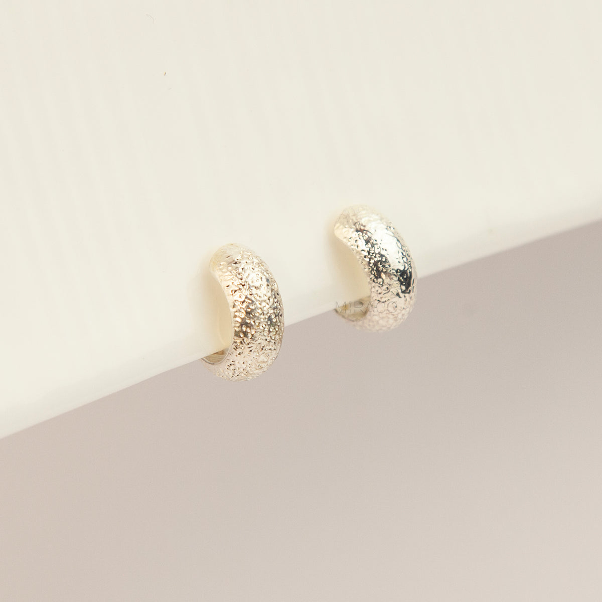 A pair of textured gold huggie hoop earrings resting on a white surface against a soft beige background.