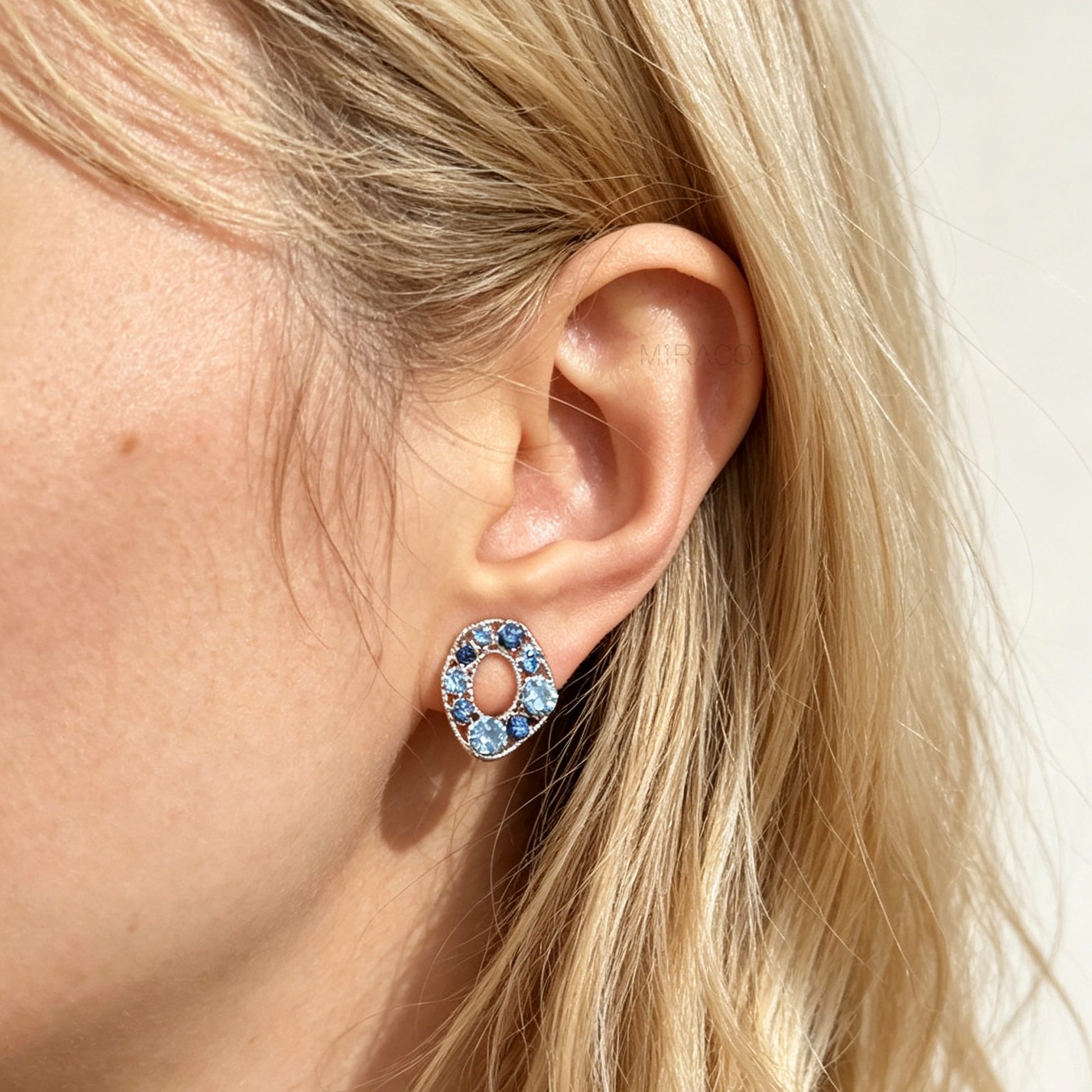 A close-up of a model's ear wearing a silver-toned oval stud earring set with light blue crystals.