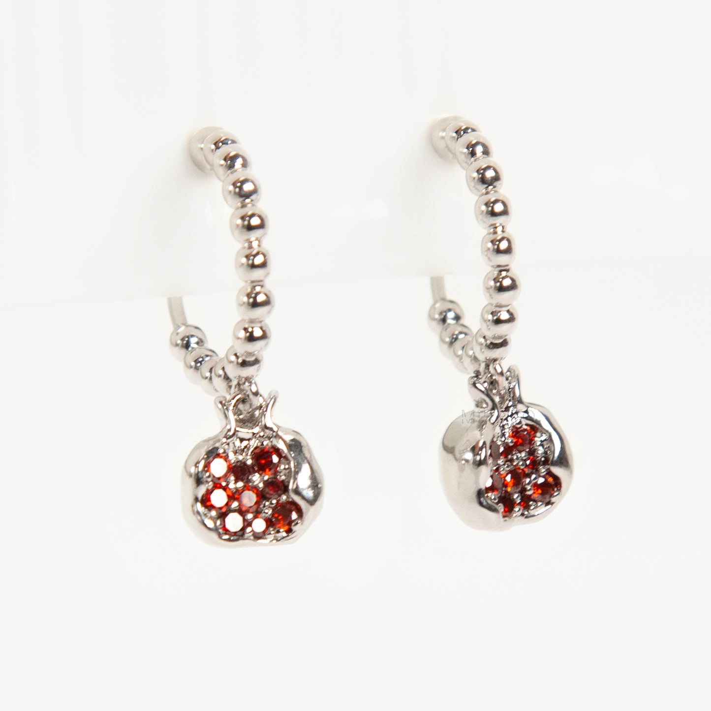 Pair of silver-tone hoop clip-on earrings with a beaded design and a red CZ-encrusted pomegranate charm dangle. Shows the Coil Back mechanism.