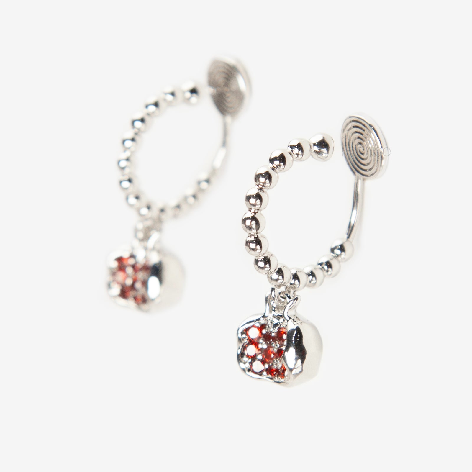 Pair of silver-tone hoop clip-on earrings with a beaded design and a red CZ-encrusted pomegranate charm dangle. Shows the Coil Back mechanism.