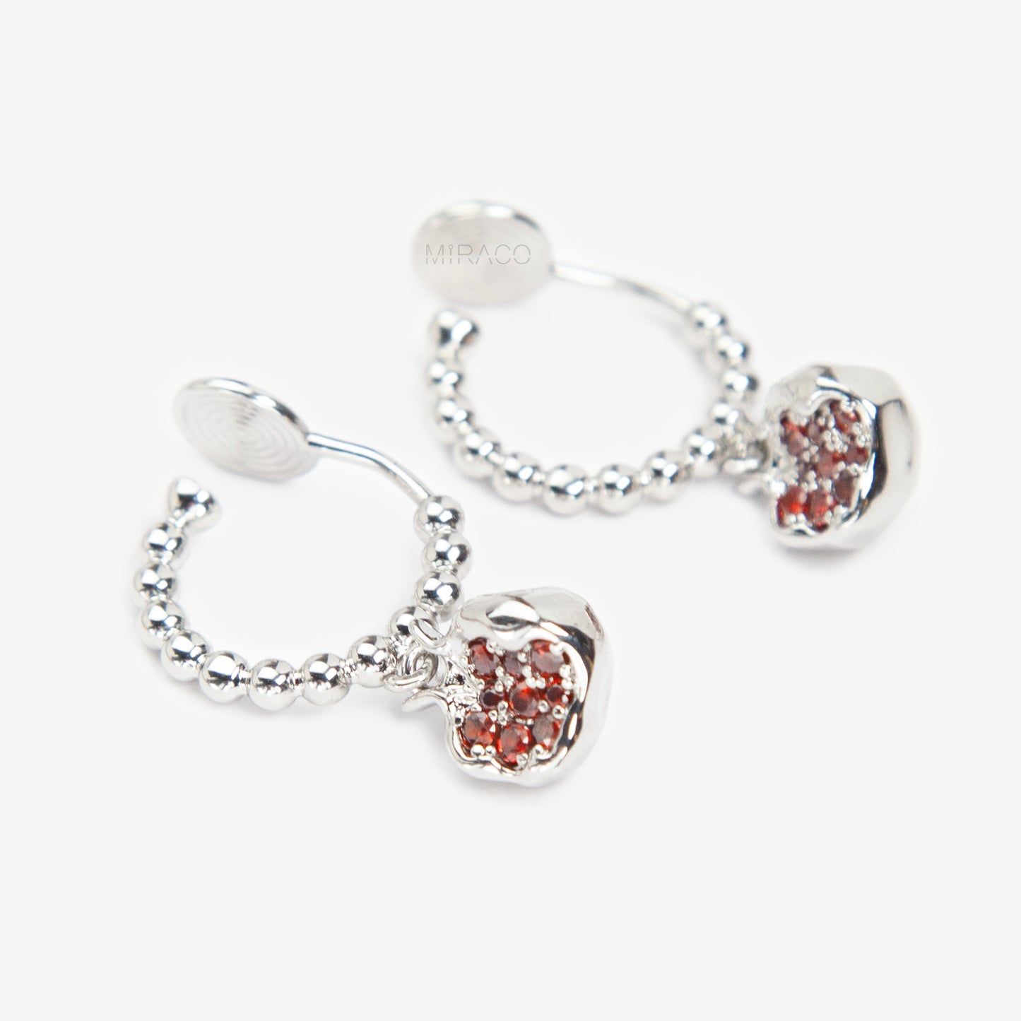 Pair of silver-tone hoop clip-on earrings with a beaded design and a red CZ-encrusted pomegranate charm dangle. Shows the Coil Back mechanism.