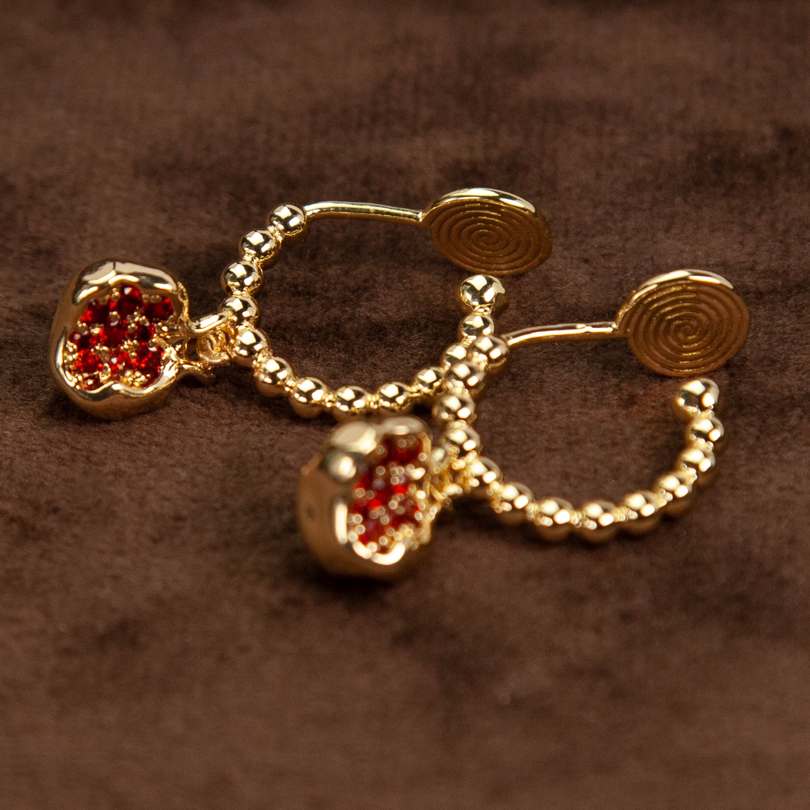 A pair of gold-tone clip-on earrings featuring a beaded drop design with a pomegranate charm filled with ruby red crystals, shown with comfortable coil backs.
