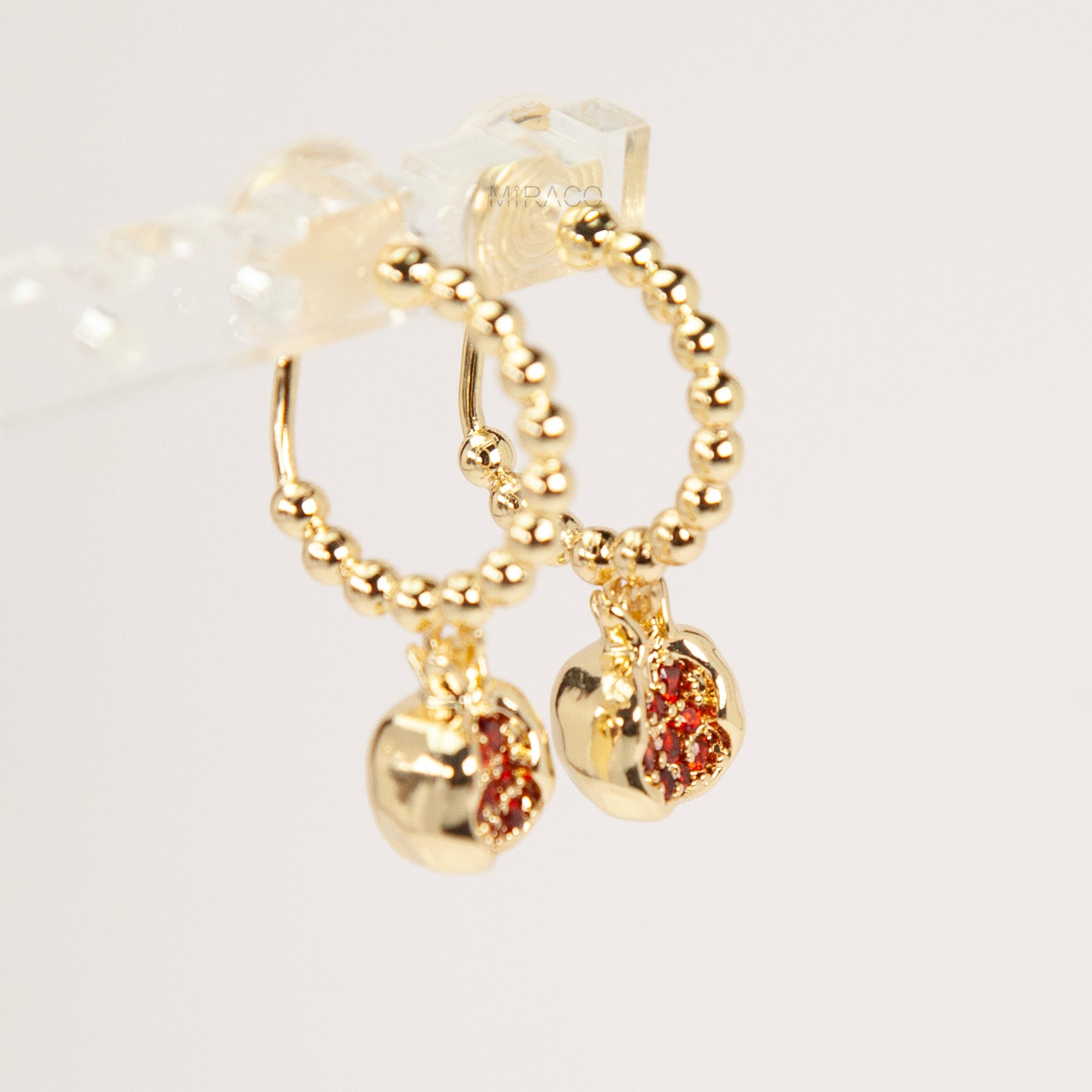 A pair of gold-tone clip-on earrings featuring a beaded drop design with a pomegranate charm filled with ruby red crystals, shown with comfortable coil backs.