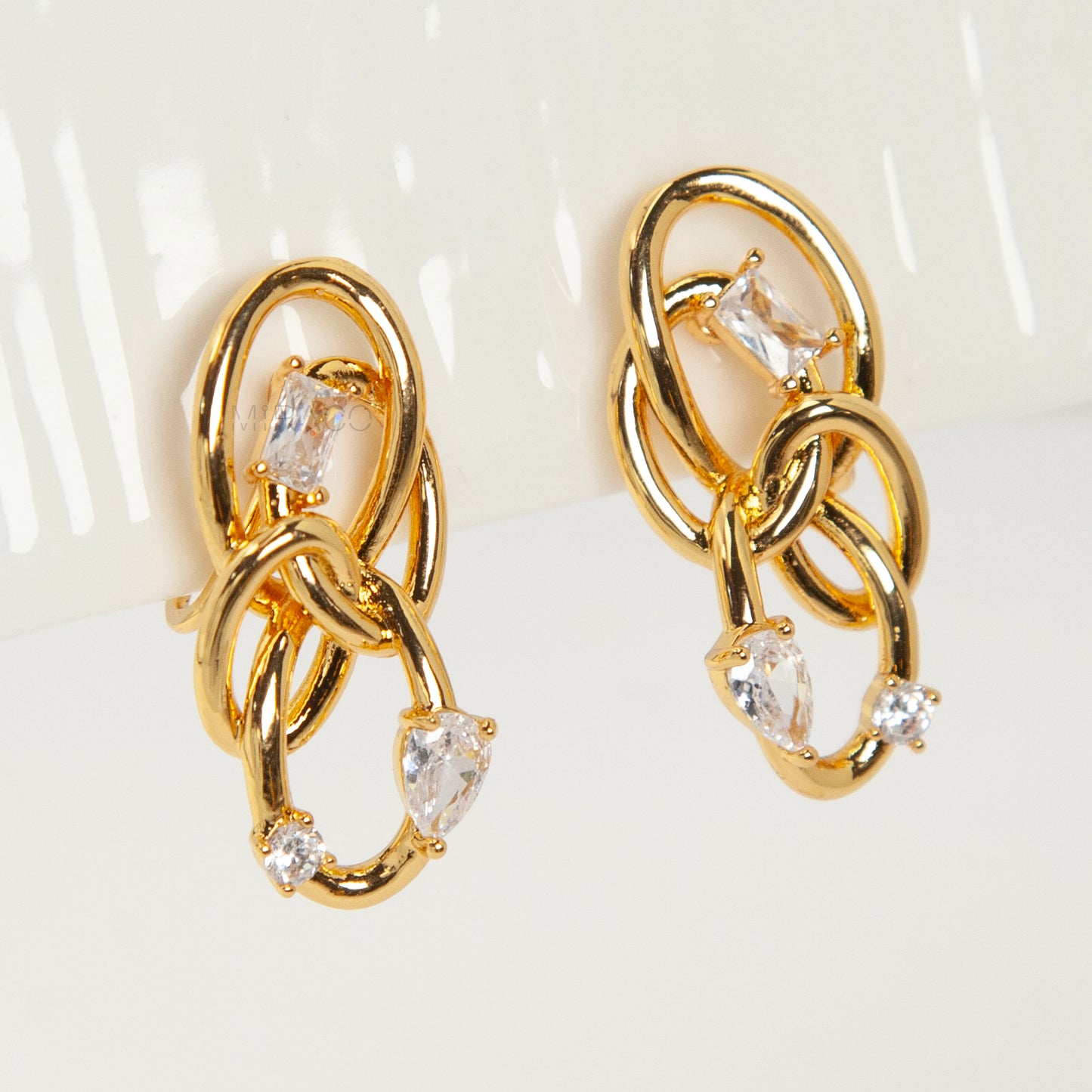 Close-up of a pair of gold interlocking double-knot clip-on earrings featuring two brilliant cut cubic zirconia stones, displayed on a white surface.