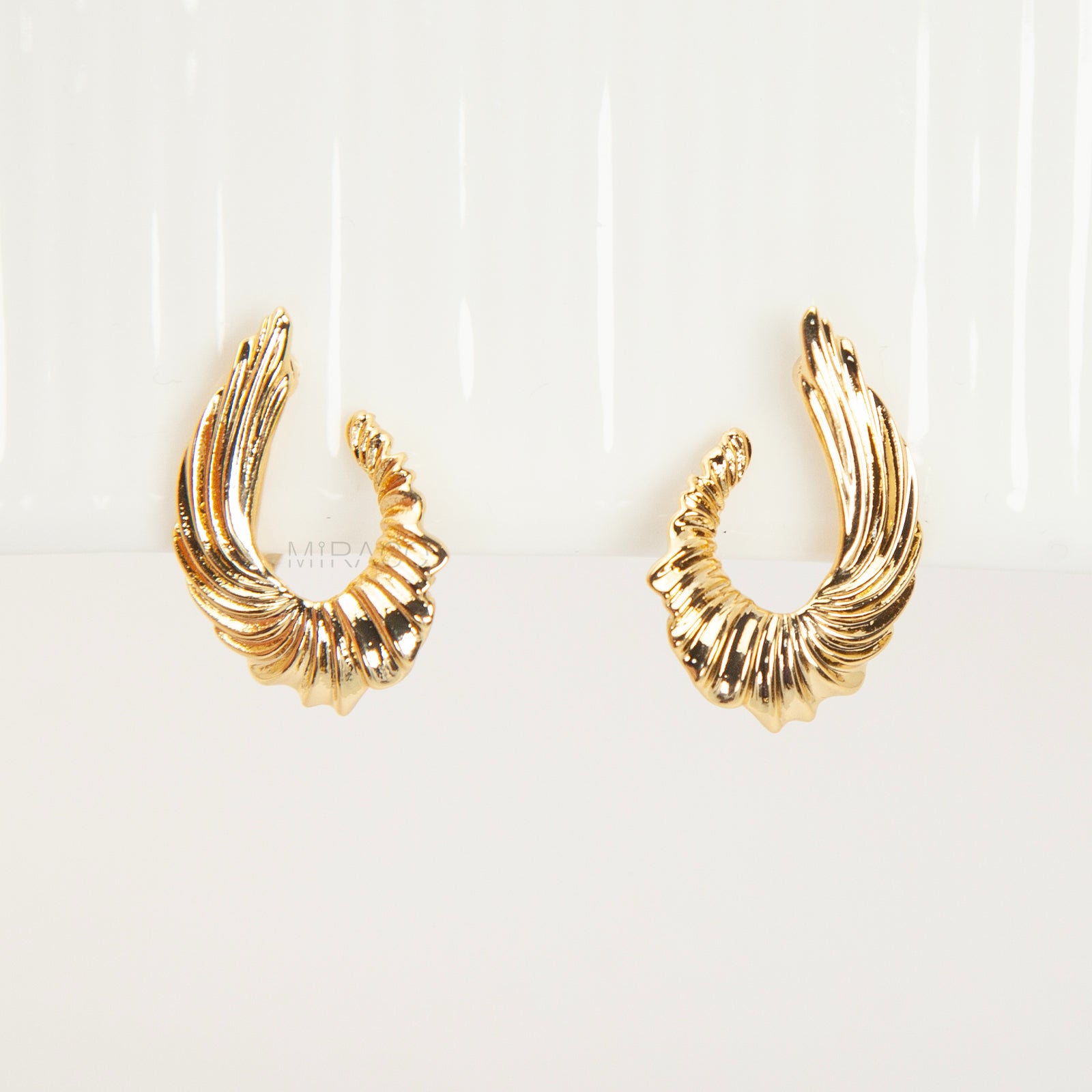 Close-up of comfortable gold-tone clip-on earrings with a ribbed, sculpted croissant or teardrop shape, worn on a model's ear. Features a secure coil back.