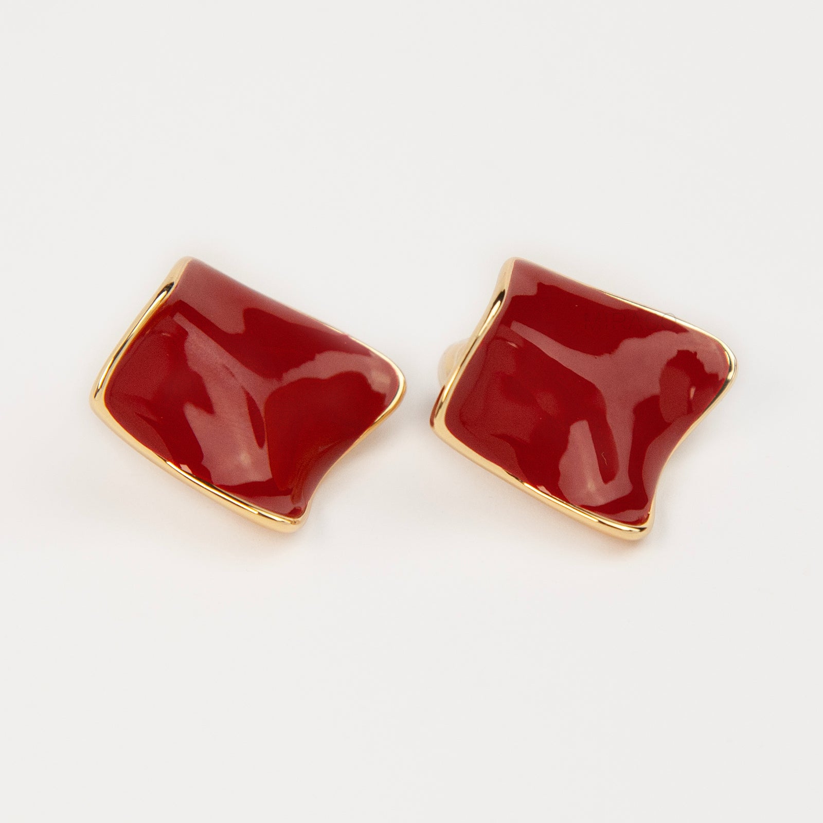 A pair of vintage-style gold clip-on earrings, showcasing a distinct wavy rhombus shape filled with smooth, glossy red enamel, fitted with comfortable, invisible-style adjustable coil backs for unpierced ears.