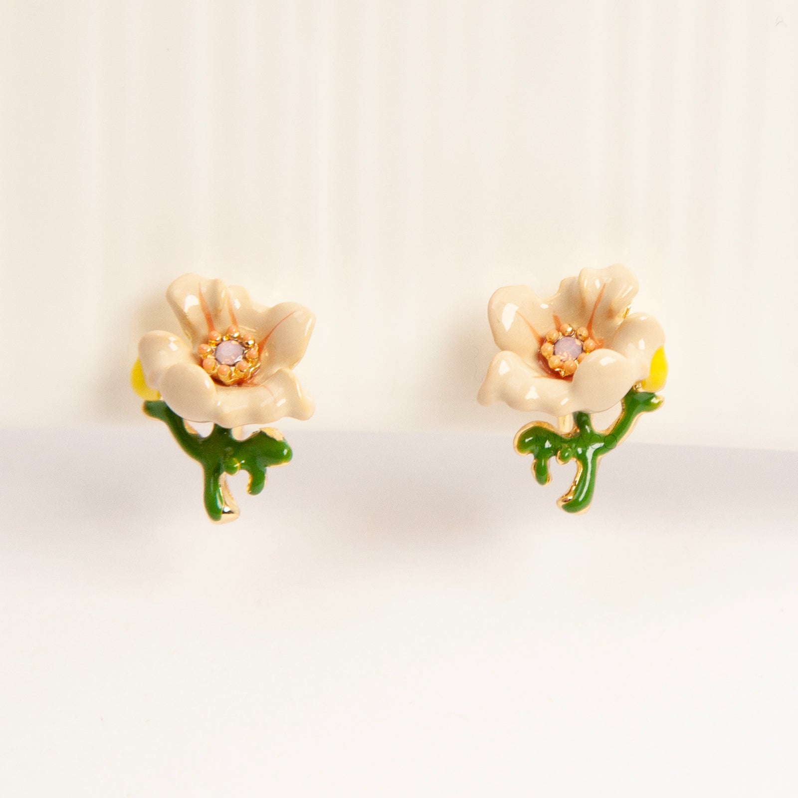 Pair of cream-colored enamel flower clip-on earrings with green stems and invisible gold-tone coil back closures on a white background.