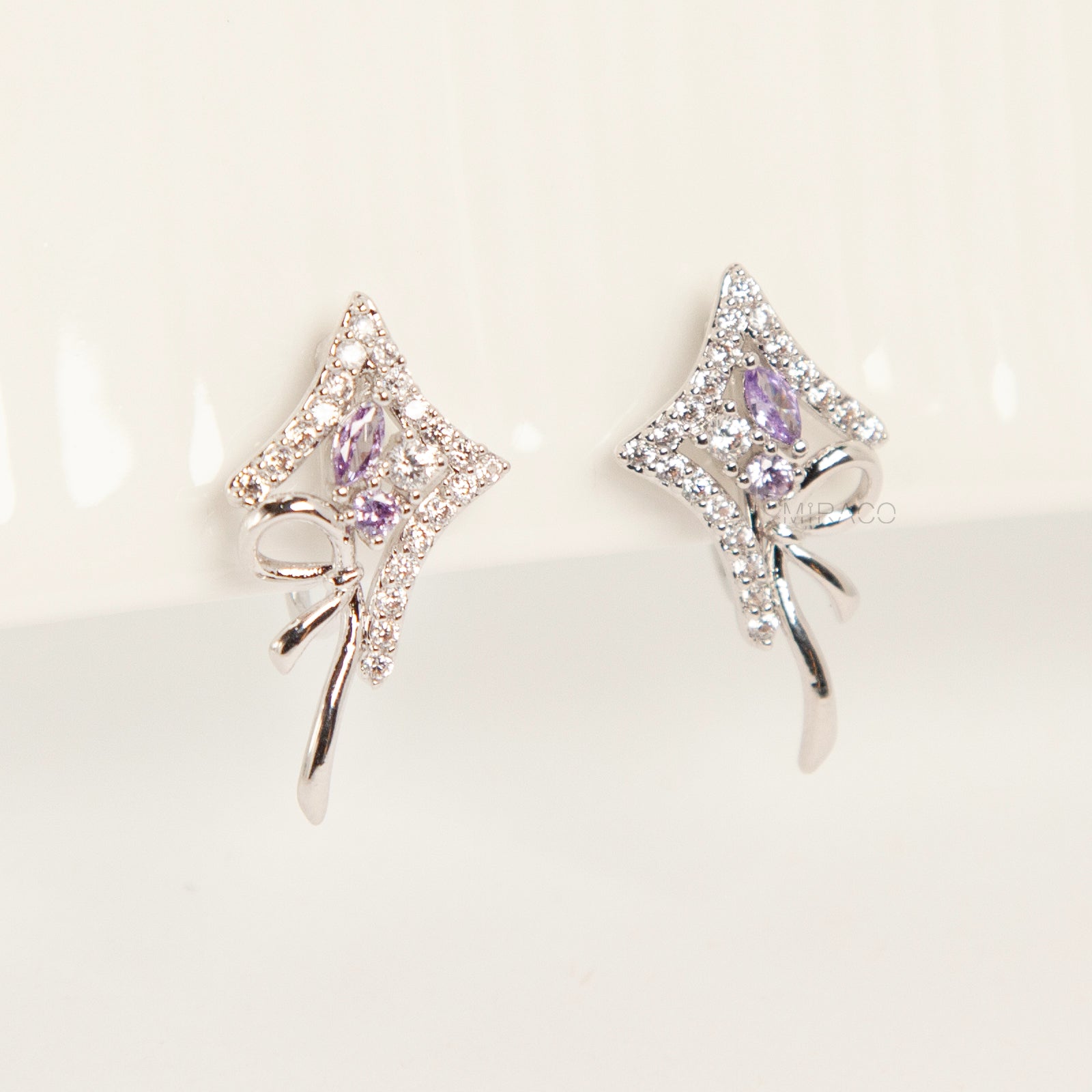 Pair of silver-tone bow-shaped clip-on earrings featuring purple amethyst stones, pave crystals, and comfortable invisible coil back closures.