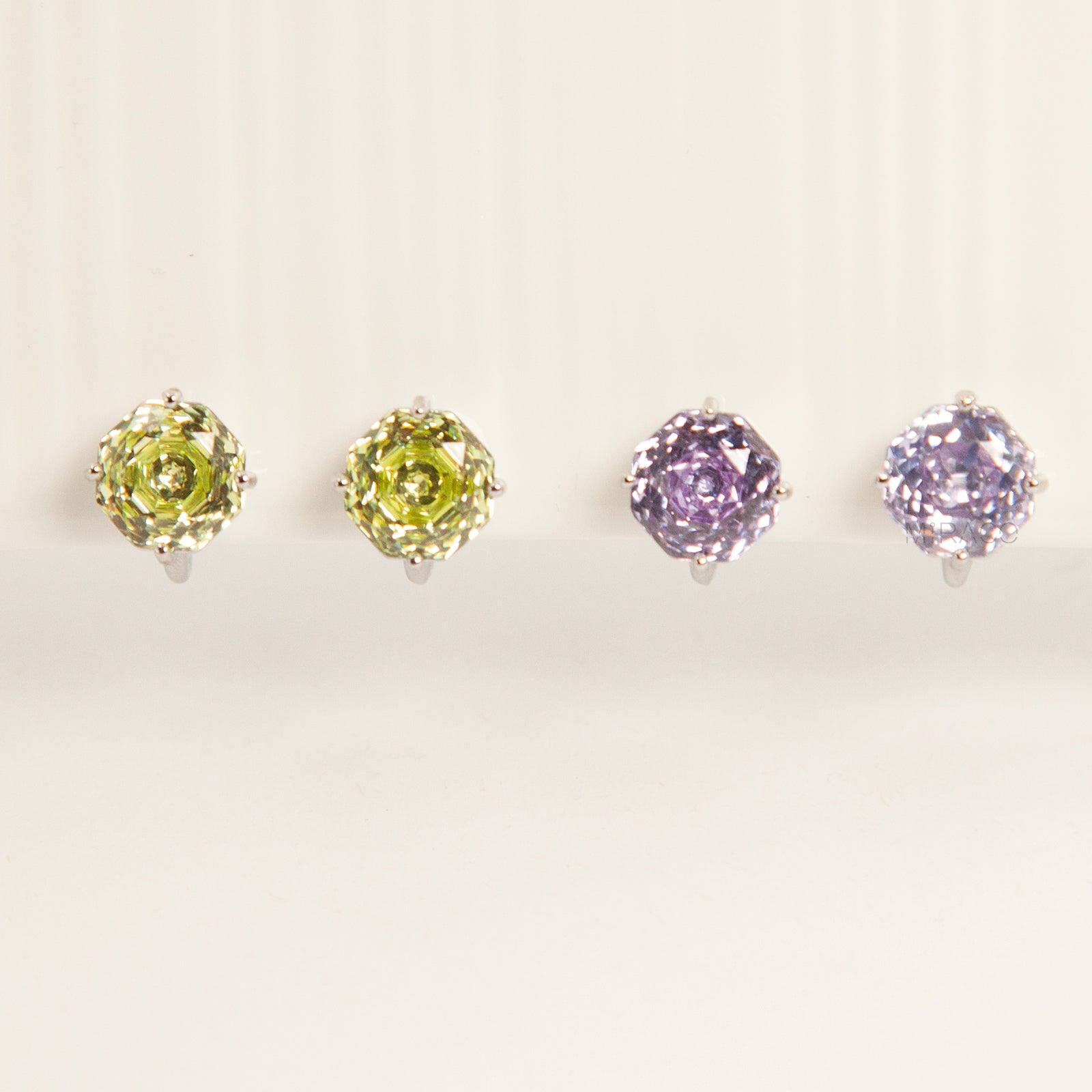 Two pairs of octagon-cut crystal clip-on stud earrings in peridot green and amethyst purple, featuring a silver-tone setting and comfortable invisible coil backs for non-pierced ears.