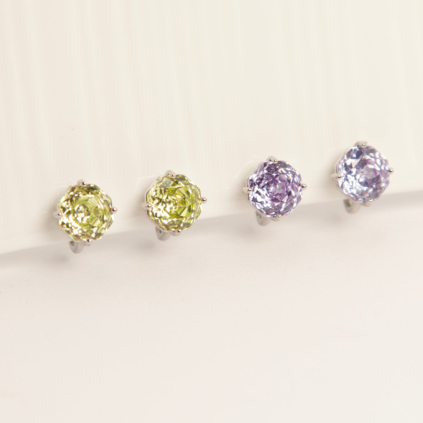 Two pairs of octagon-cut crystal clip-on stud earrings in peridot green and amethyst purple, featuring a silver-tone setting and comfortable invisible coil backs for non-pierced ears.