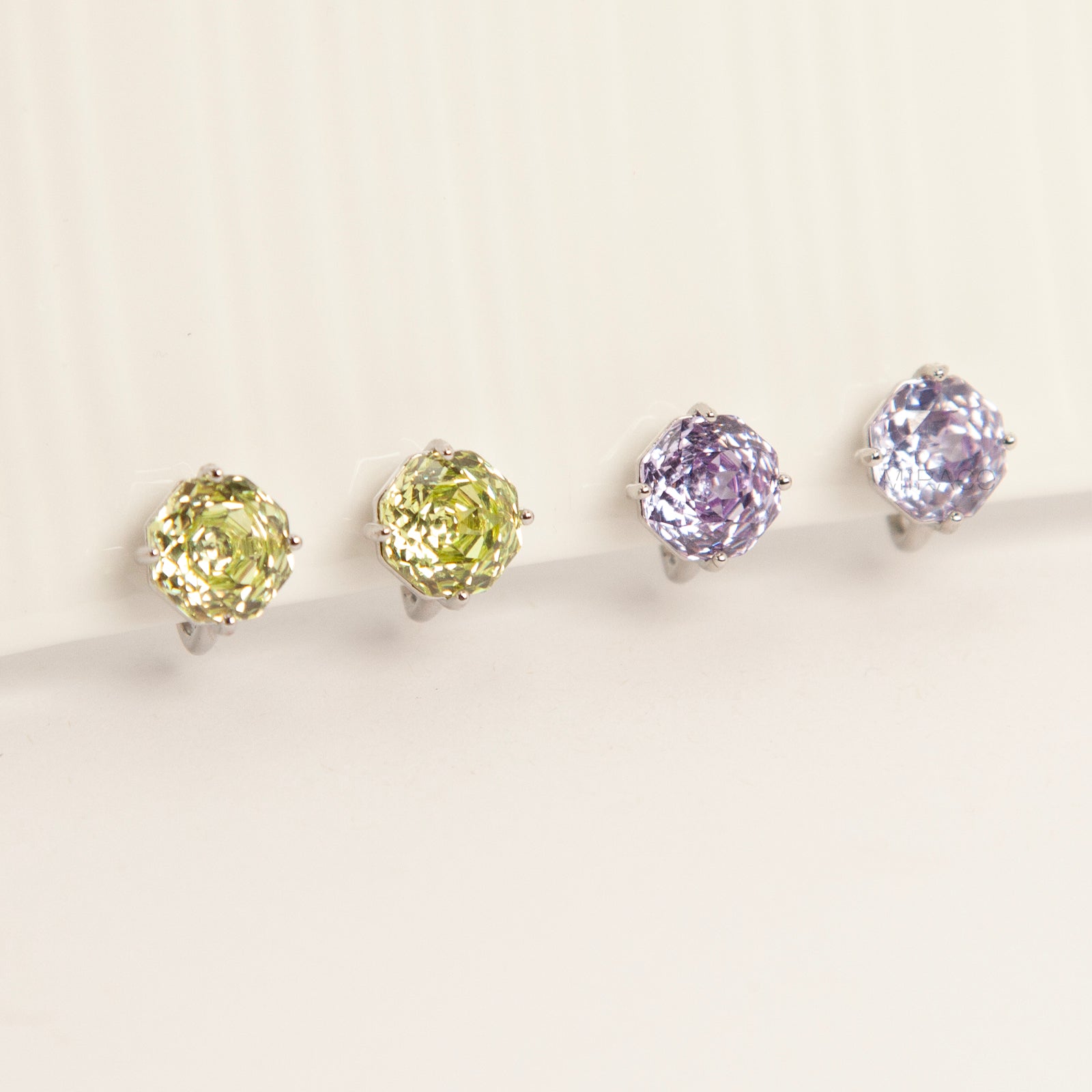 Two pairs of octagon-cut crystal clip-on stud earrings in peridot green and amethyst purple, featuring a silver-tone setting and comfortable invisible coil backs for non-pierced ears.