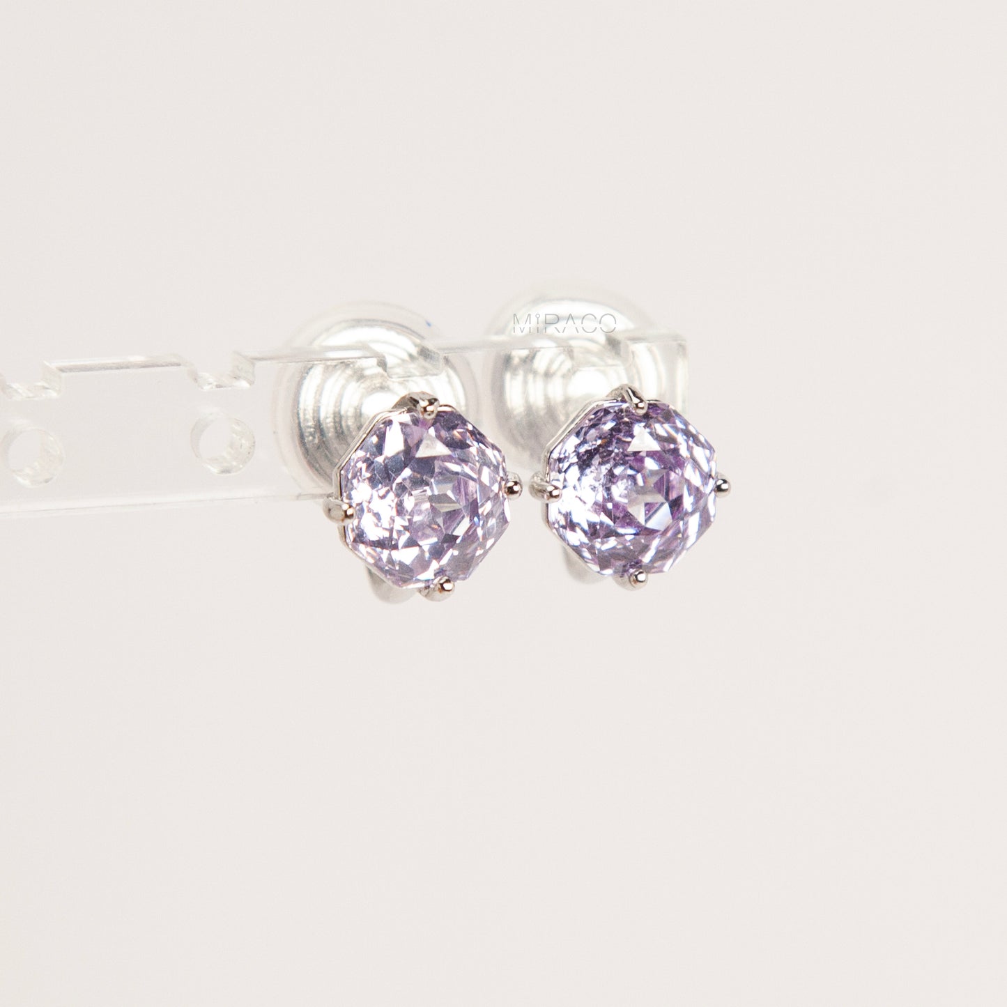 Two pairs of octagon-cut crystal clip-on stud earrings in peridot green and amethyst purple, featuring a silver-tone setting and comfortable invisible coil backs for non-pierced ears.