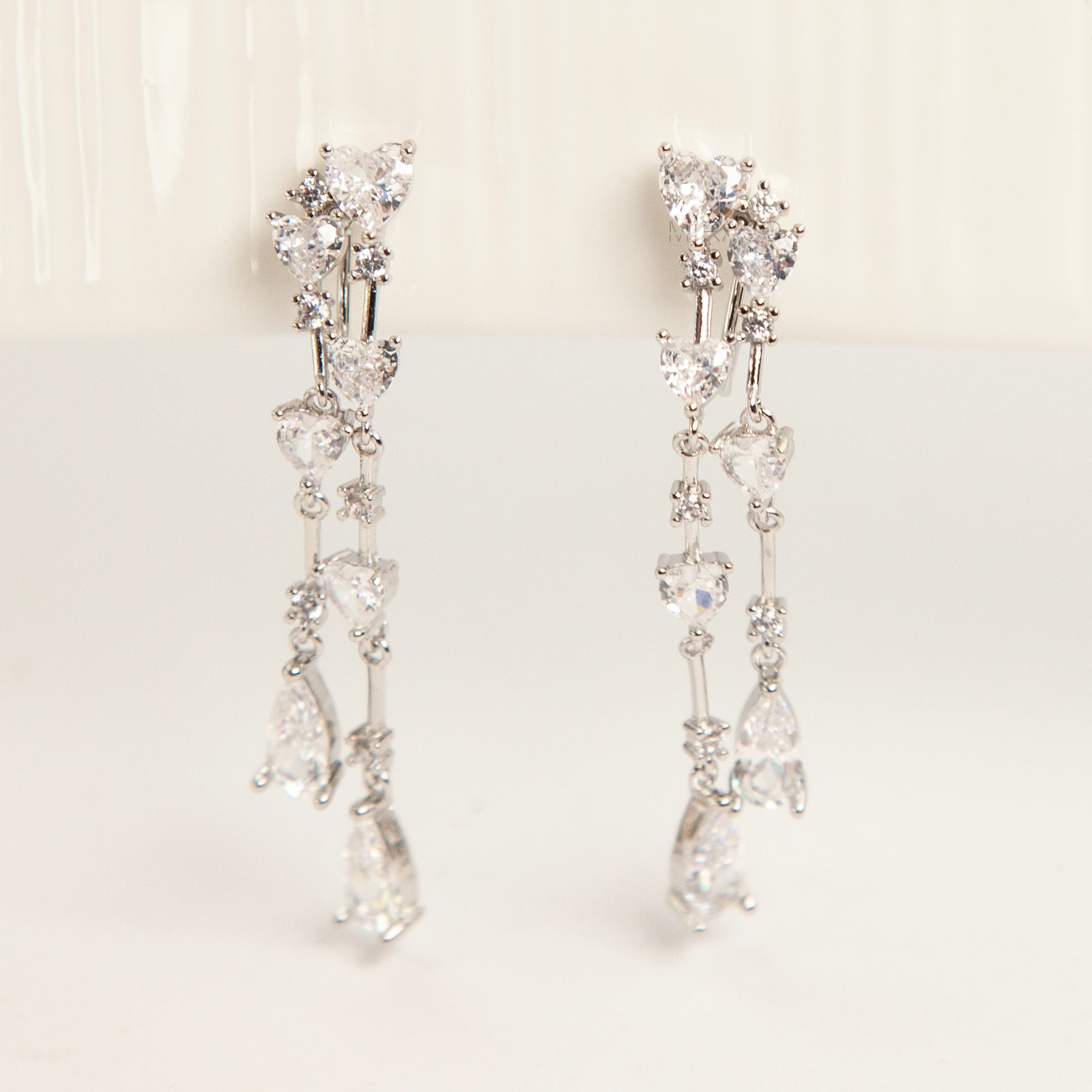 Pair of silver-toned cascading clip-on earrings featuring heart-shaped and teardrop cubic zirconia crystals on a dainty drop chain with a comfortable coil back.