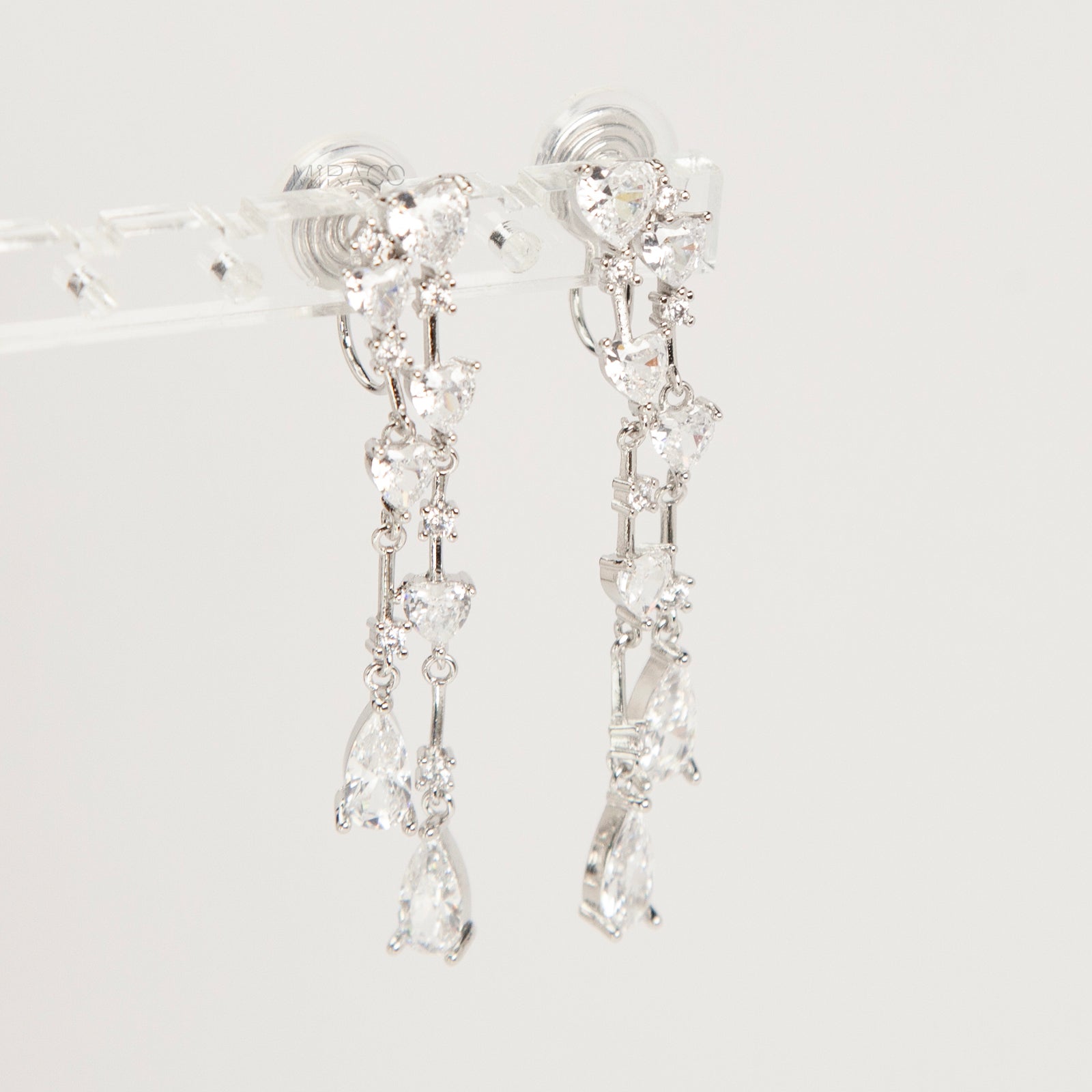Pair of silver-toned cascading clip-on earrings featuring heart-shaped and teardrop cubic zirconia crystals on a dainty drop chain with a comfortable coil back.
