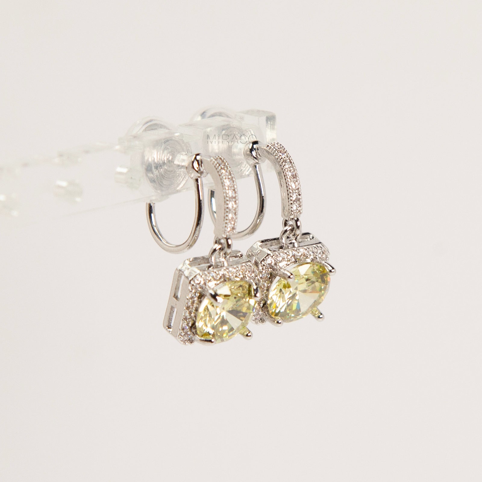 Side view of silver-toned clip-on earrings showing the adjustable invisible coil back mechanism and sparkling olive green cushion-cut drop stones.