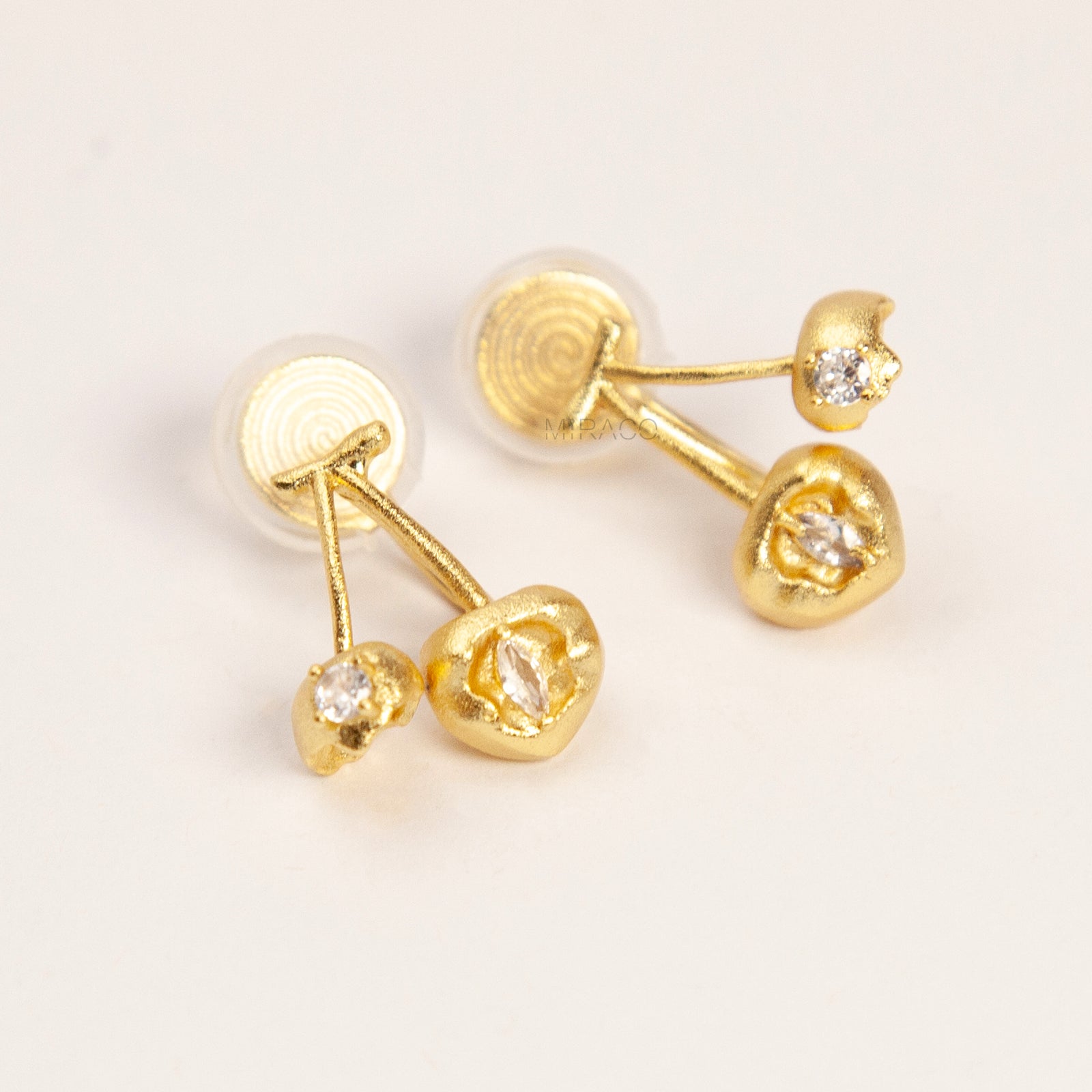A pair of matte gold cherry-shaped clip-on earrings with small embedded crystals, featuring gold-tone adjustable coil-spring backs for comfortable wear.