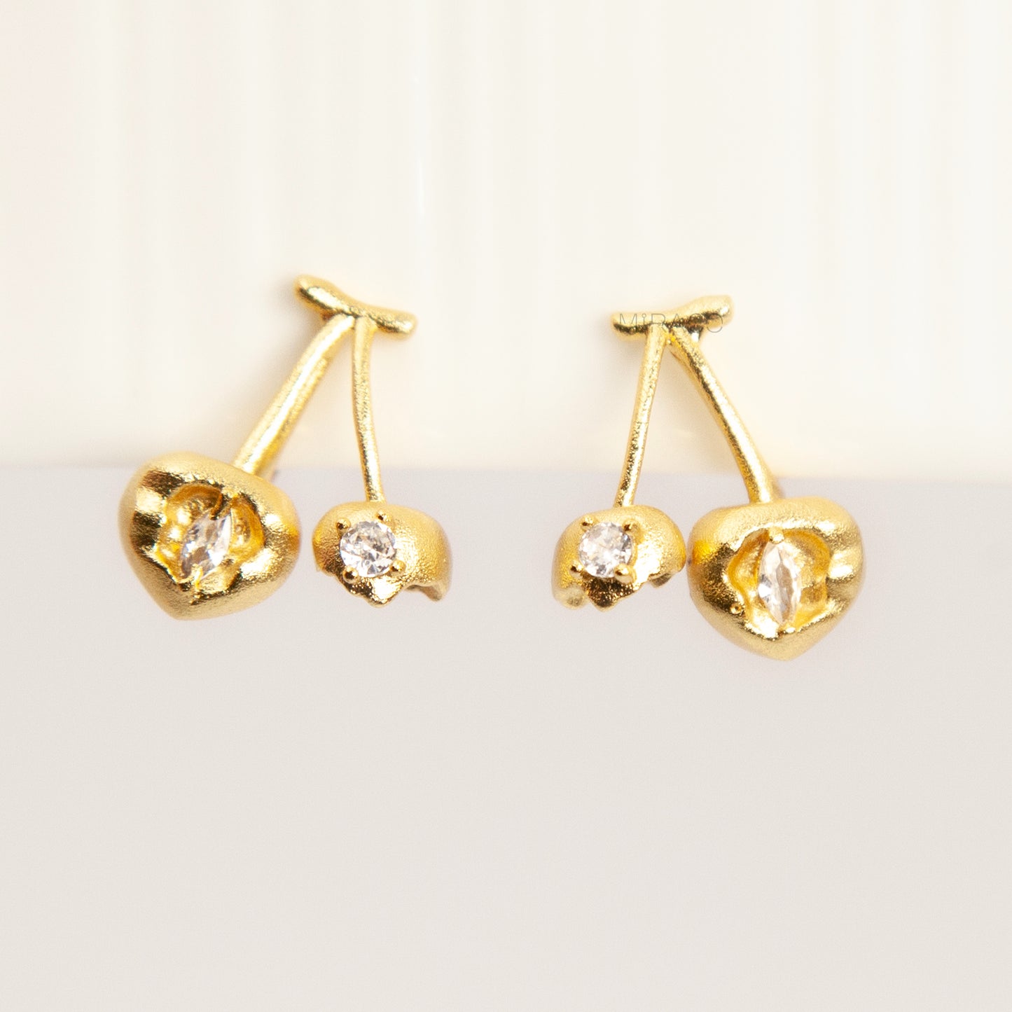 A pair of matte gold cherry-shaped clip-on earrings with small embedded crystals, featuring gold-tone adjustable coil-spring backs for comfortable wear.