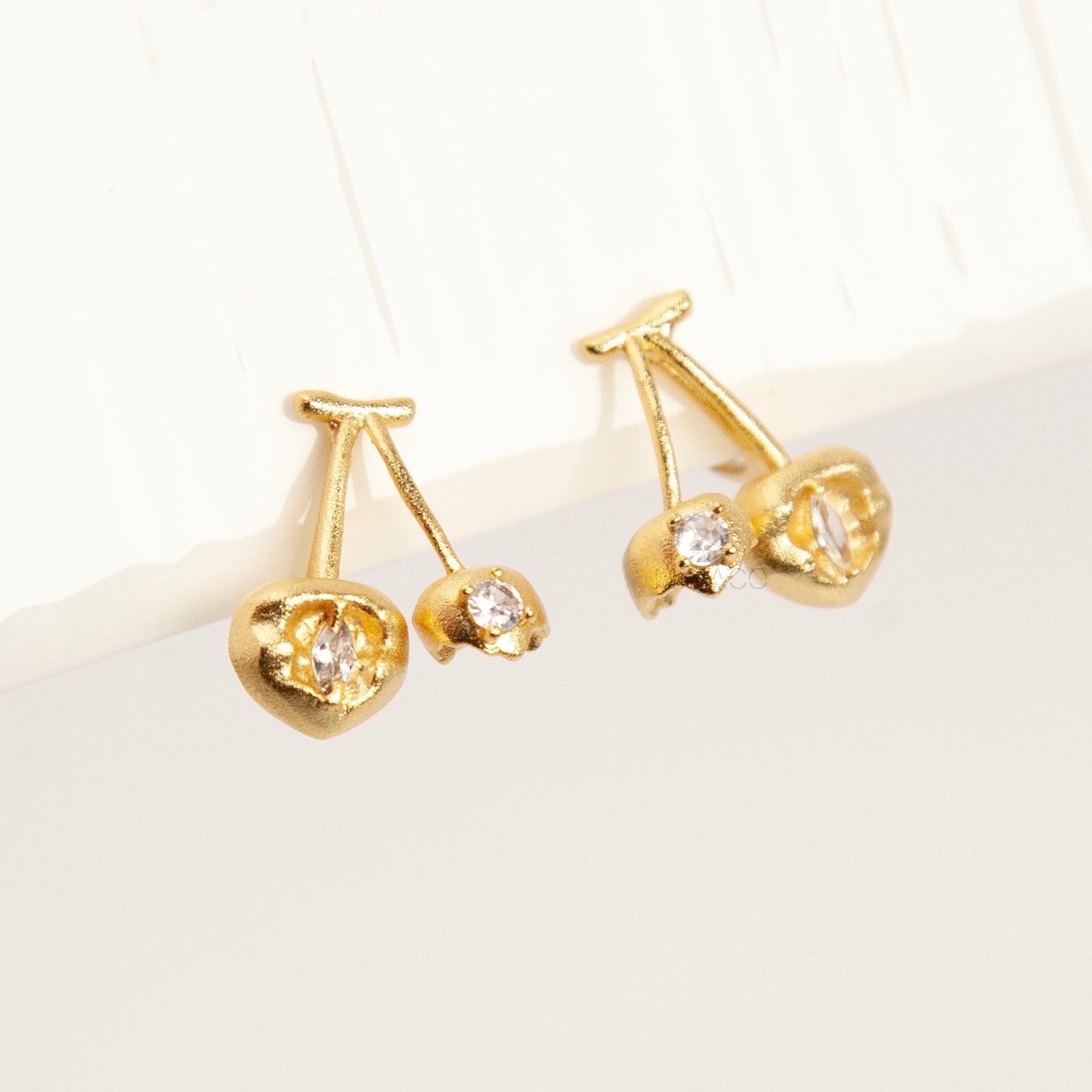 A pair of matte gold cherry-shaped clip-on earrings with small embedded crystals, featuring gold-tone adjustable coil-spring backs for comfortable wear.