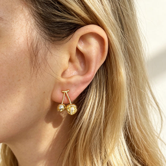 A pair of matte gold cherry-shaped clip-on earrings with small embedded crystals, featuring gold-tone adjustable coil-spring backs for comfortable wear.