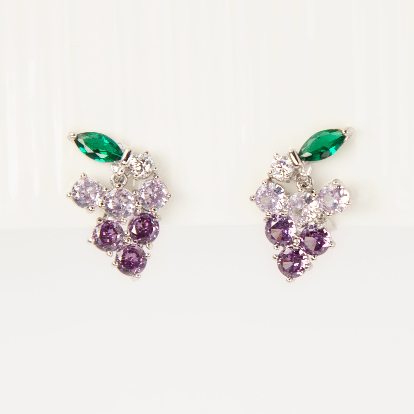 A pair of silver-tone clip-on earrings in the shape of a grape cluster, featuring various shades of round purple crystals and a green marquise-cut crystal leaf