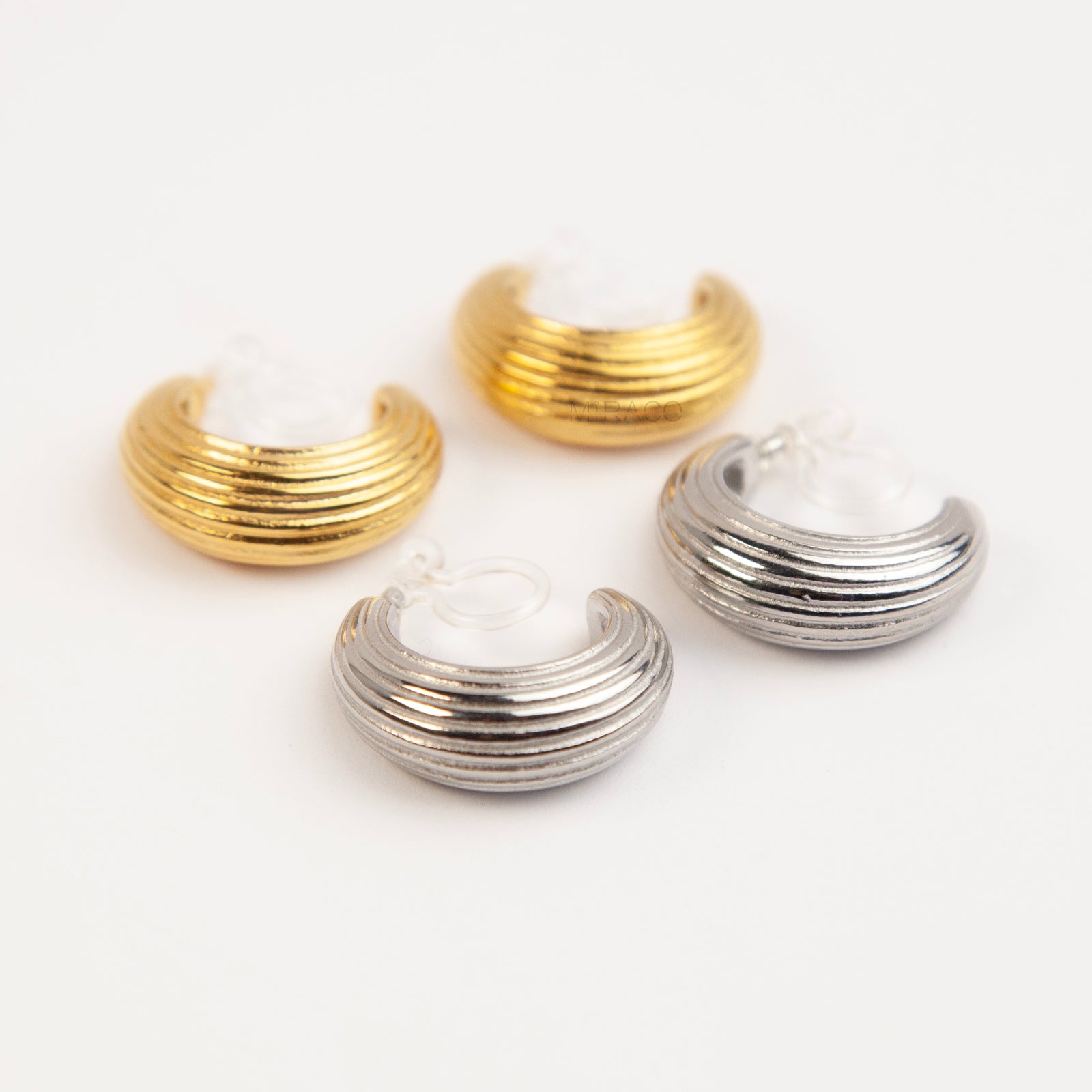 Two pairs of ribbed C-shaped hoop earrings, one in gold and one in silver, featuring clear resin clip-on backs for non-pierced ears