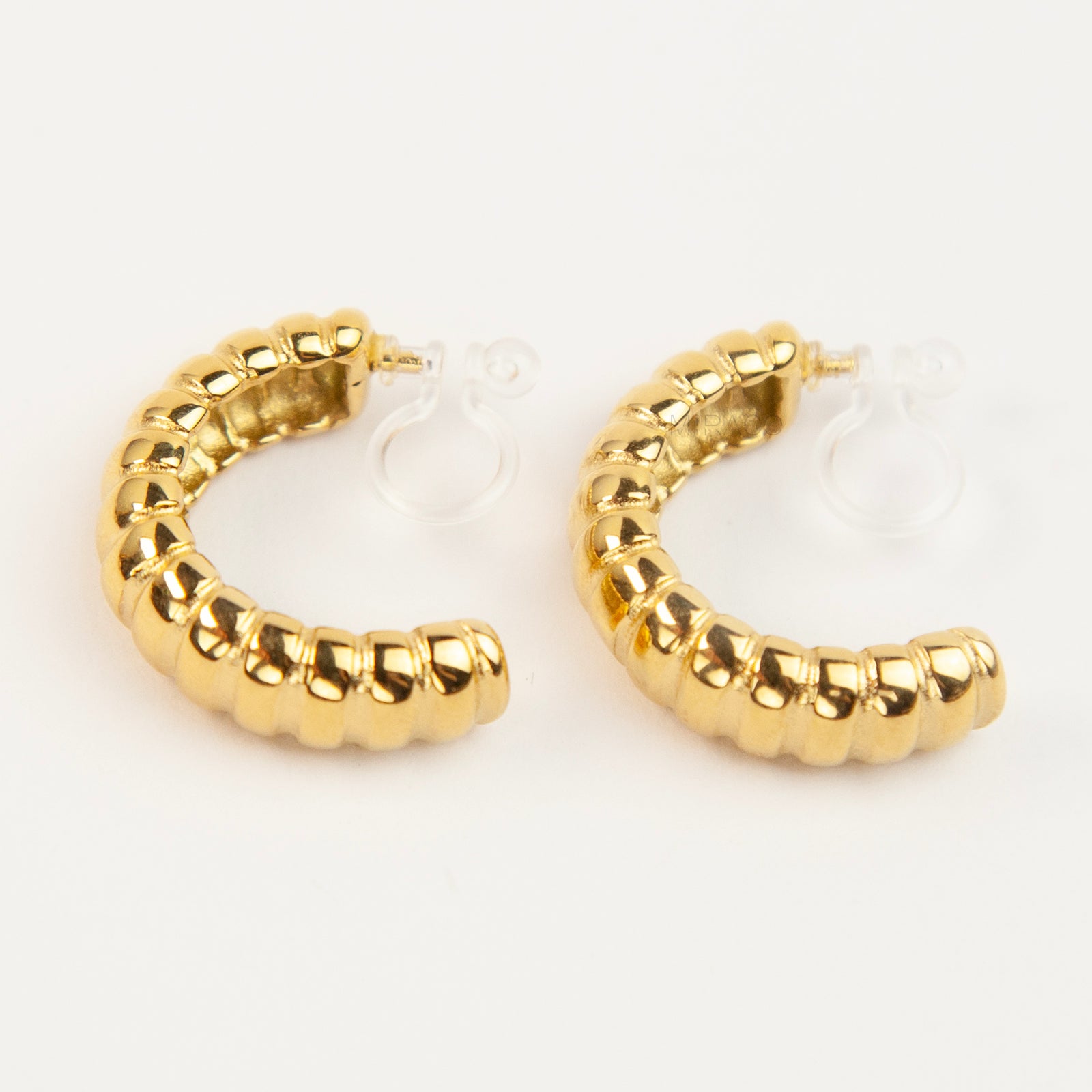 Two pairs of textured croissant-style C-hoop earrings, one pair in gold and one pair in silver, featuring clear invisible resin clip-on backs