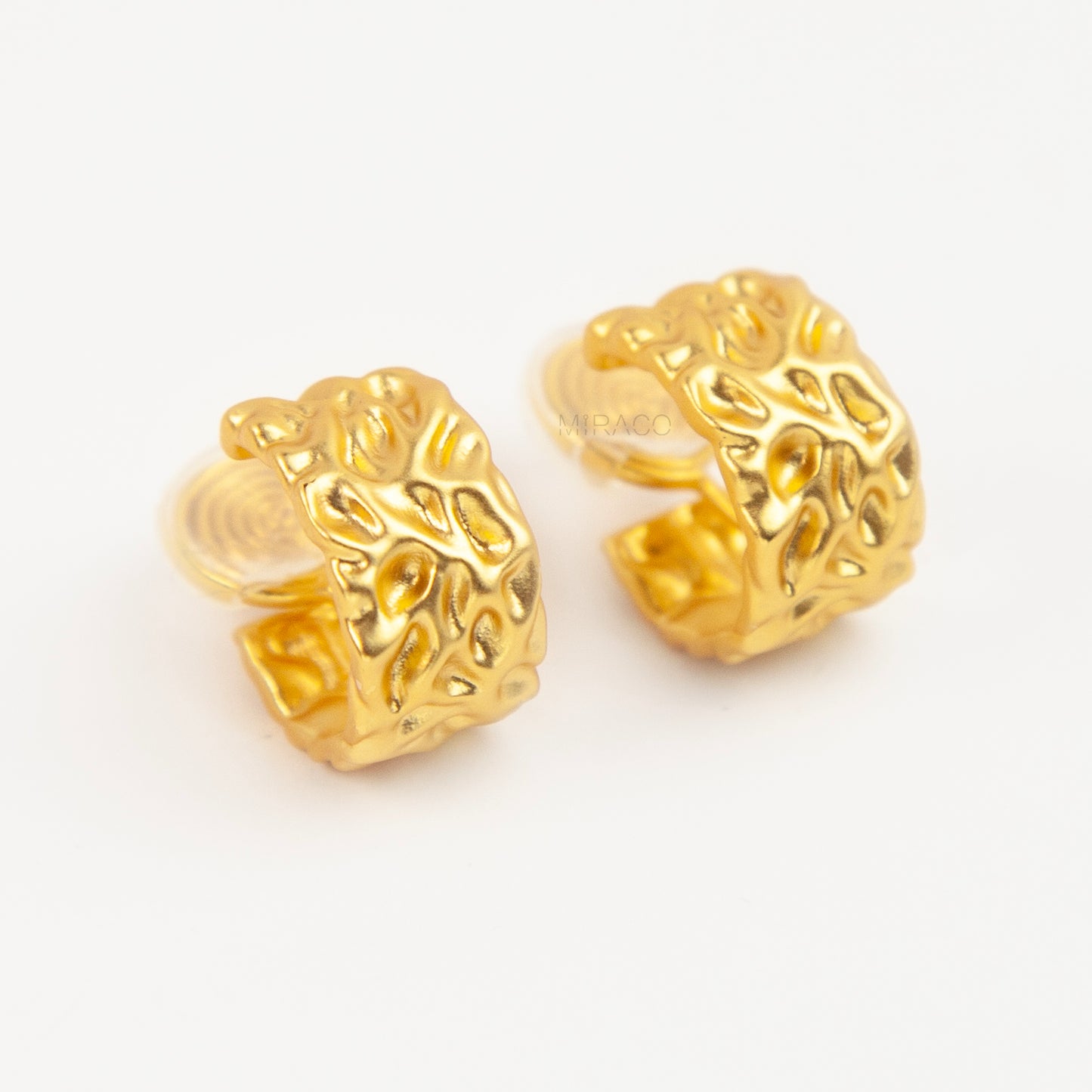 A pair of wide, matte gold-tone clip-on hoop earrings with an organic hammered molten texture, featuring gold adjustable coil-spring backs