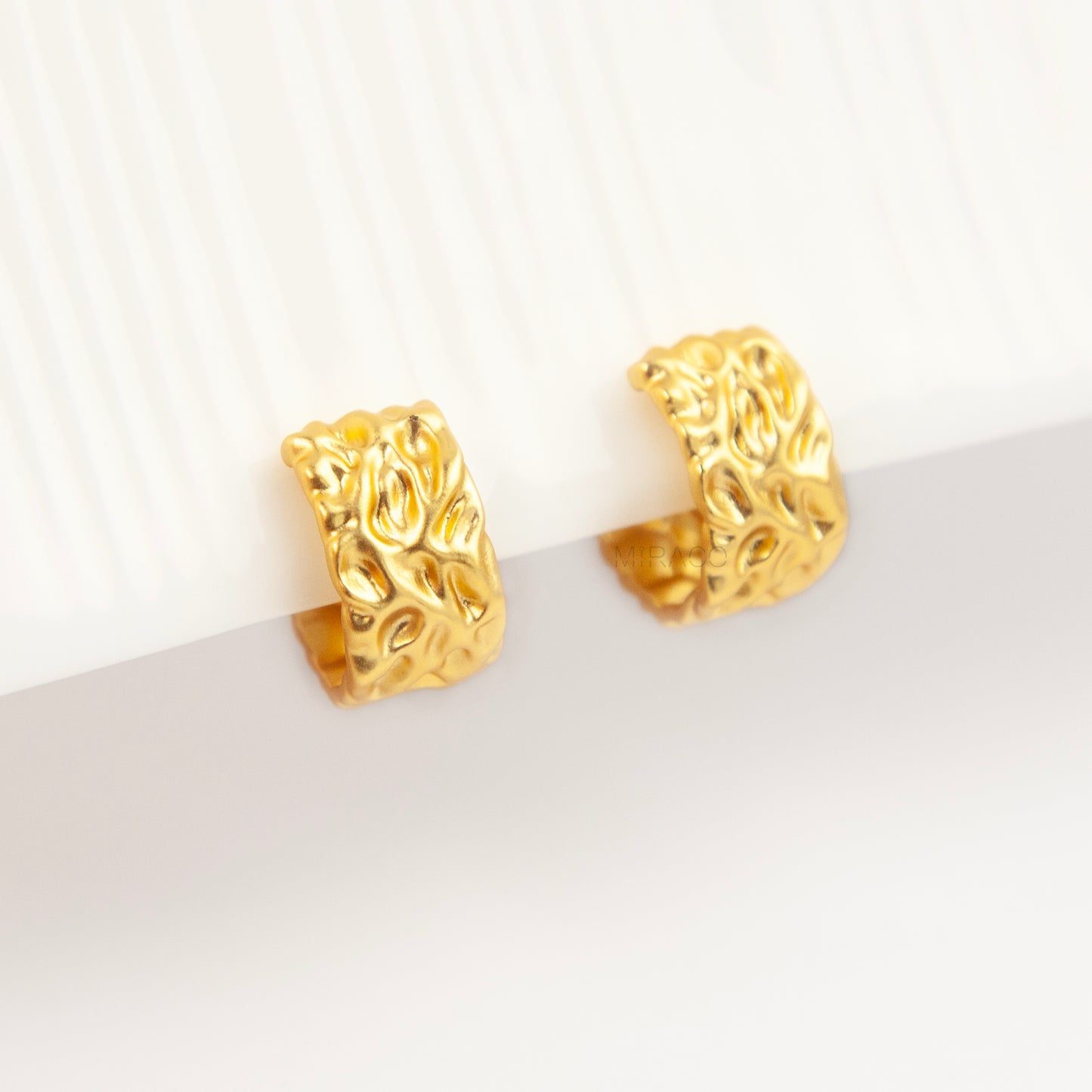 A pair of wide, matte gold-tone clip-on hoop earrings with an organic hammered molten texture, featuring gold adjustable coil-spring backs