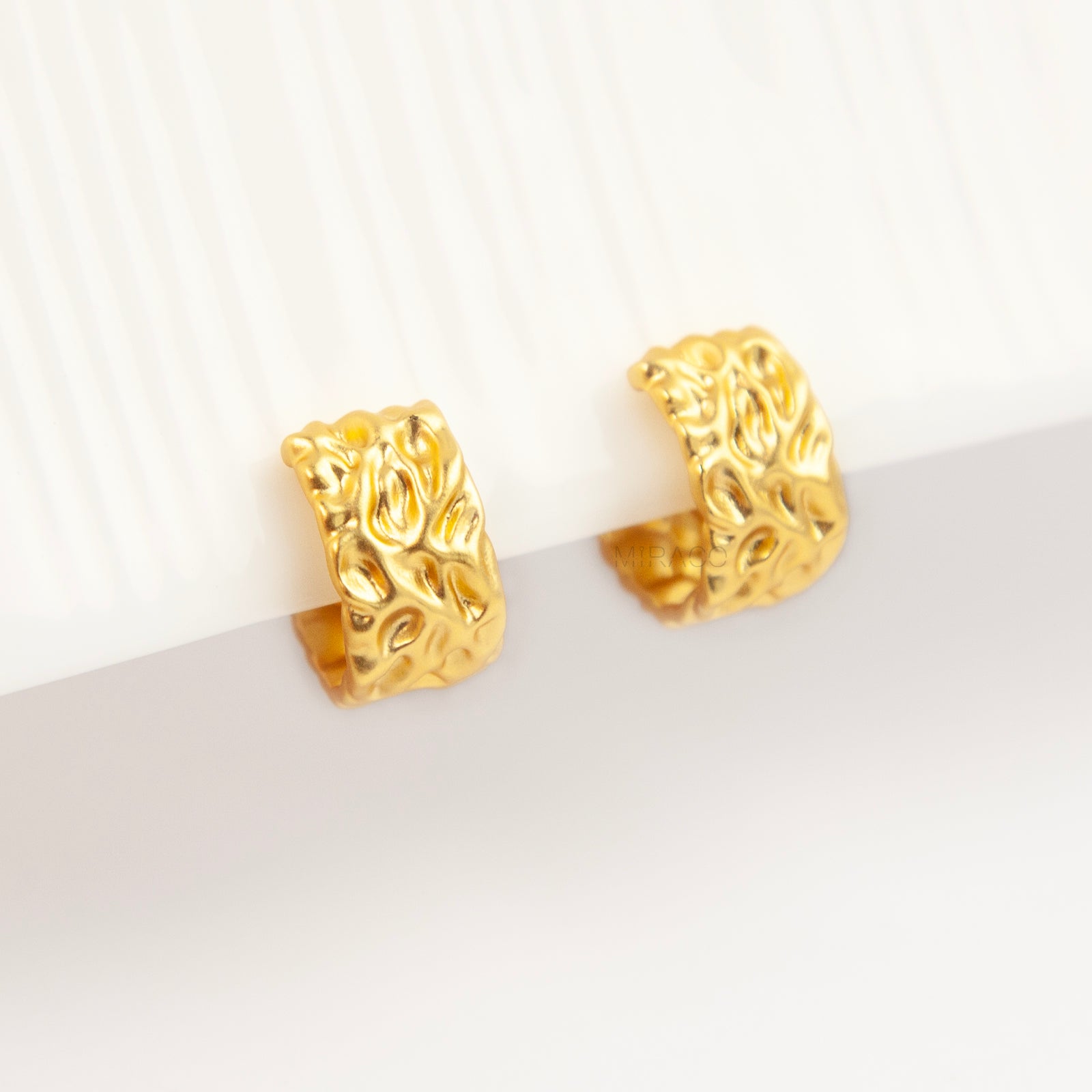 A pair of wide, matte gold-tone clip-on hoop earrings with an organic hammered molten texture, featuring gold adjustable coil-spring backs