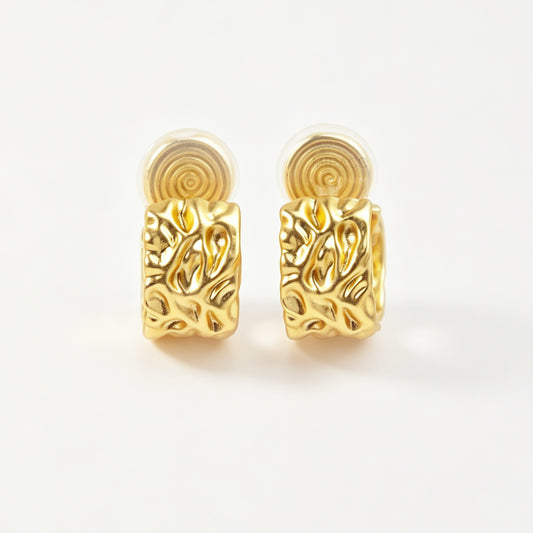 A pair of wide, matte gold-tone clip-on hoop earrings with an organic hammered molten texture, featuring gold adjustable coil-spring backs