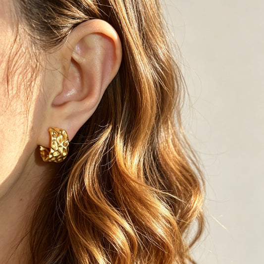 A pair of wide, matte gold-tone clip-on hoop earrings with an organic hammered molten texture, featuring gold adjustable coil-spring backs