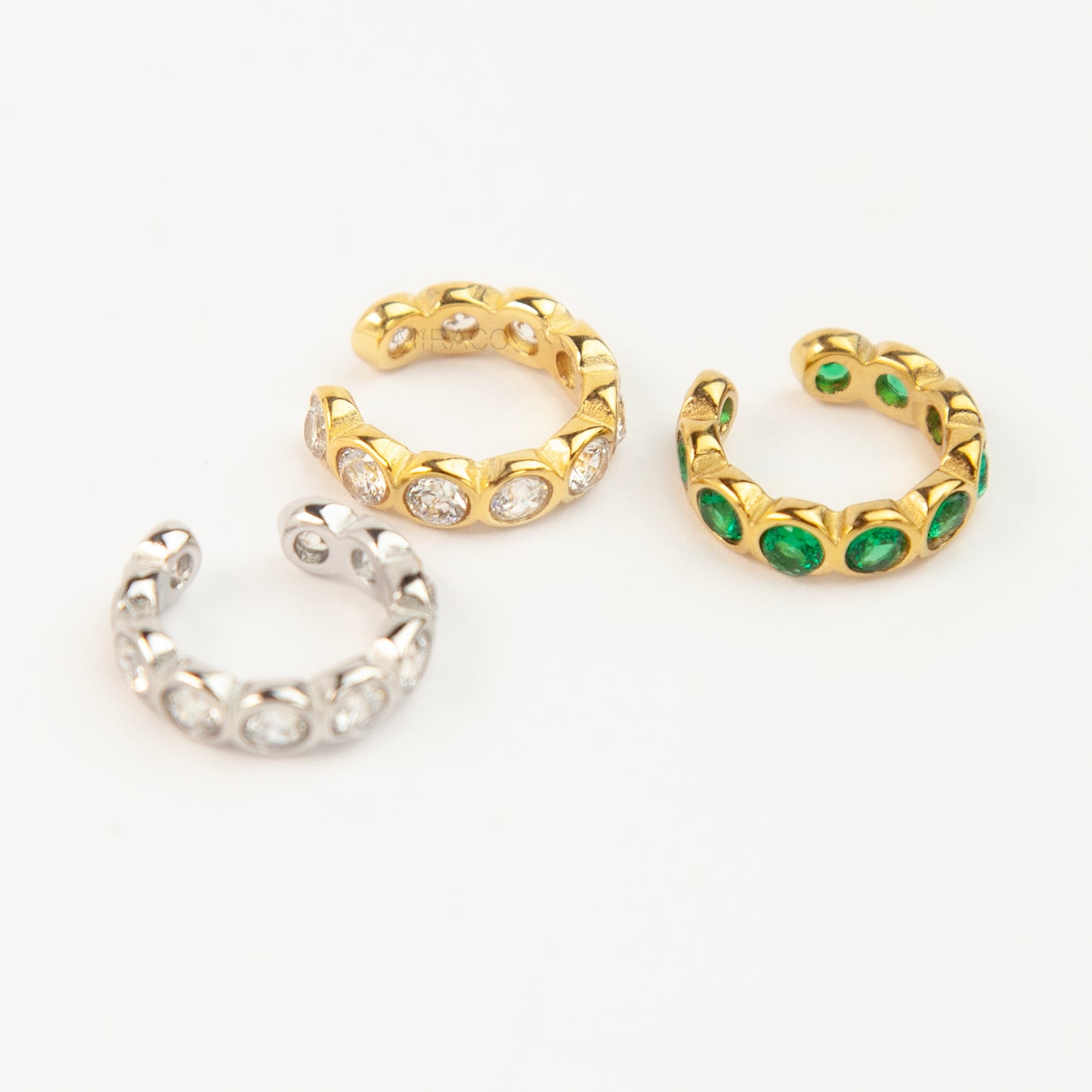 Three single eternity ear cuffs with bezel-set stones, including one silver with clear crystals, one gold with clear crystals, and one gold with emerald green crystals.
