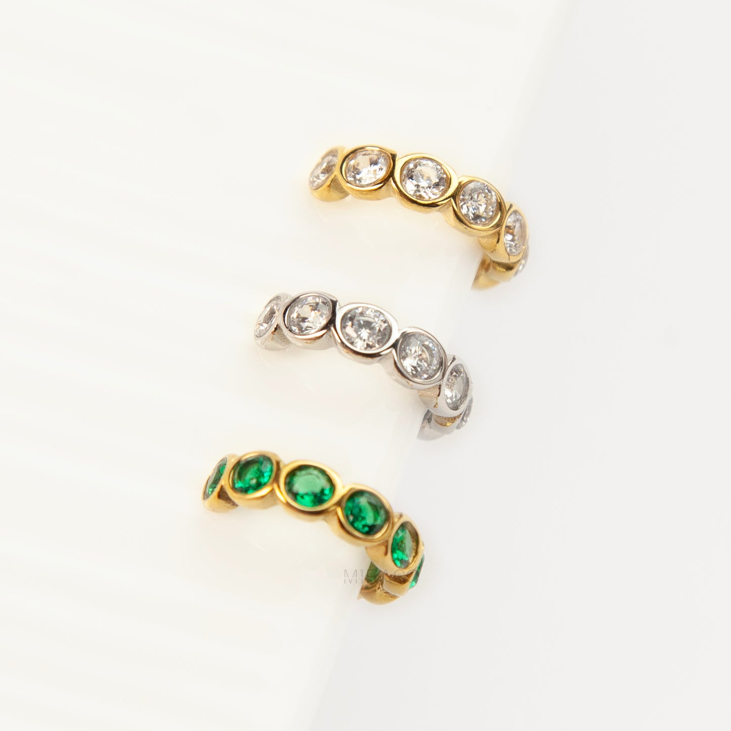 Three single eternity ear cuffs with bezel-set stones, including one silver with clear crystals, one gold with clear crystals, and one gold with emerald green crystals.